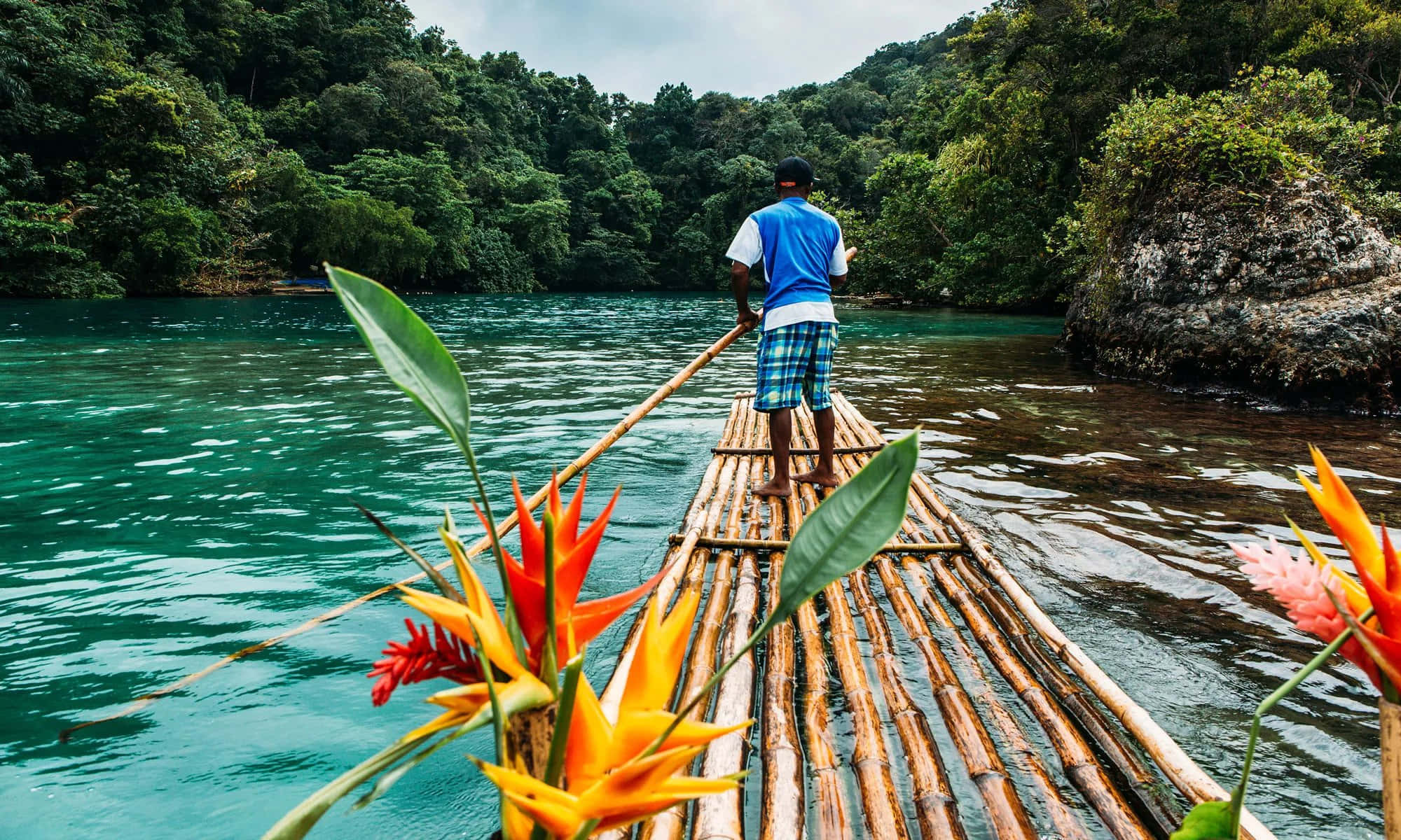 Jamaica 桌布 壁紙 — 一名男子划著竹筏穿過碧綠色的水域，四周環繞著鮮豔的橙色花朵和茂密的綠色叢林。