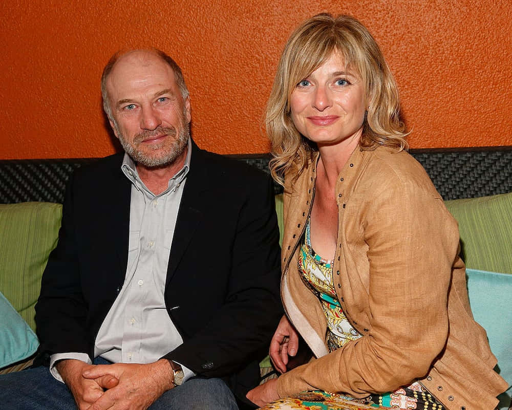 Ted Levine 桌布 壁紙 — 一名男子和一名女子並排坐在梳化上，背景是帶有紋理的橙色牆壁，並有綠色和藍色靠墊。