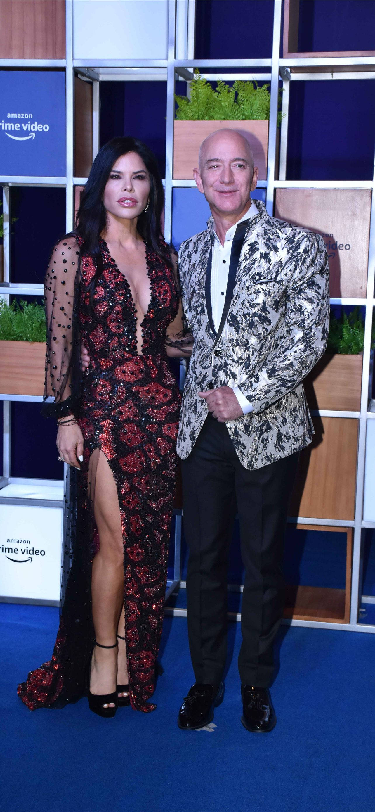 Bezos 桌布 壁紙 — 一名男子和一名女子在亞馬遜Prime Video品牌背景前，優雅地站在藍色地毯上，背景有幾何面板和綠色植物。
