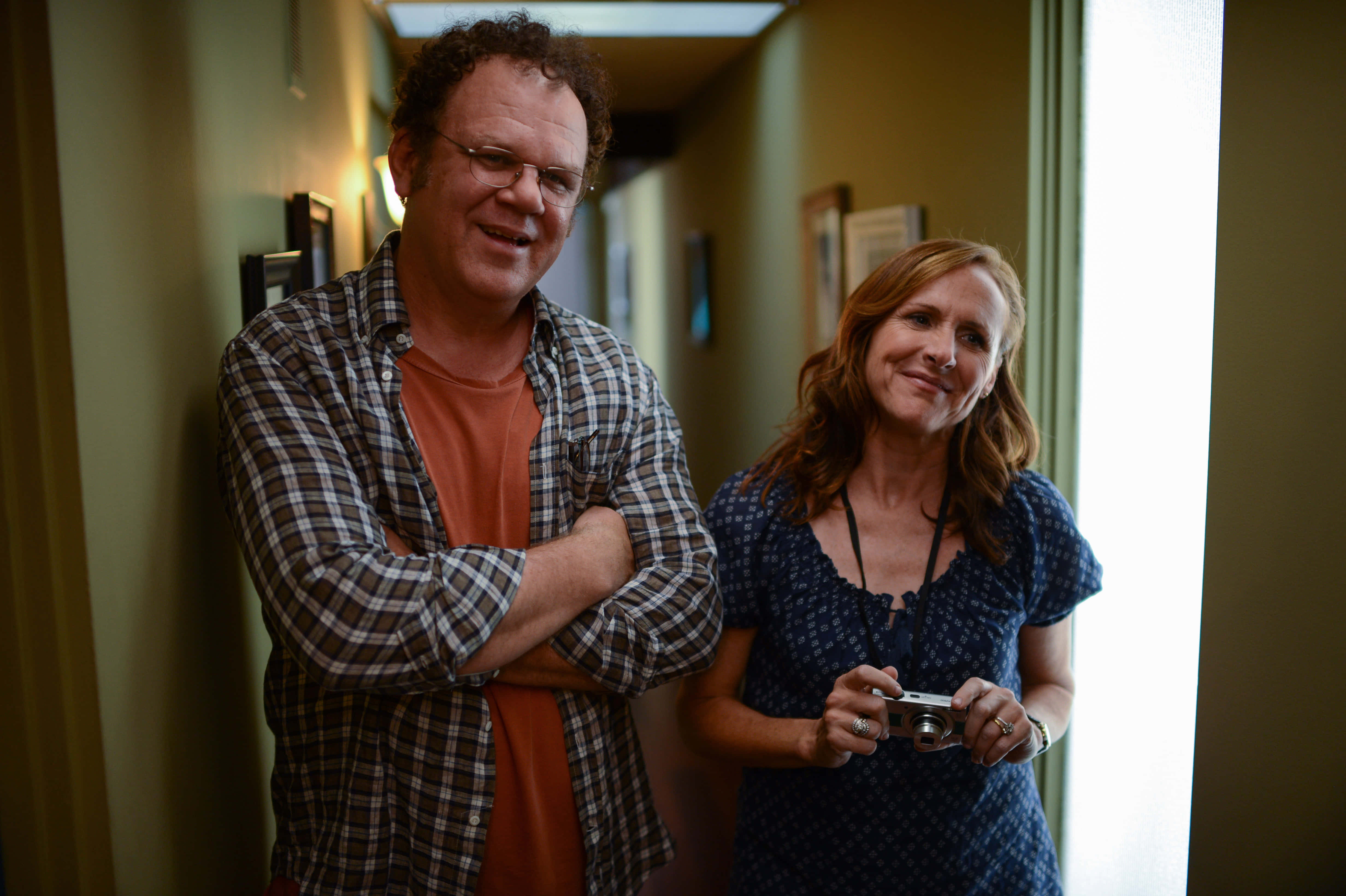 John C. Reilly 桌布 壁紙 — 一名男子和一名女子在陽光照射的走廊中溫暖地微笑，女子手持相機。