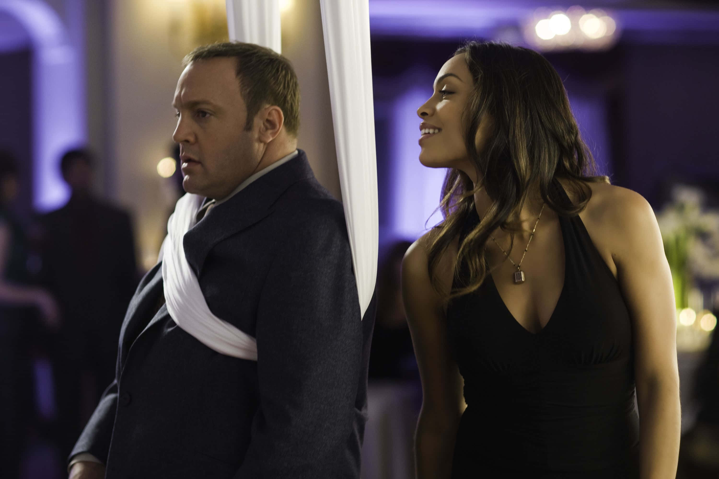 Kevin James 桌布 壁紙 — 一名男子和一名女子穿著優雅的正式服裝，在燈光昏暗、帶有紫色調的派對中背對背站立，周圍有柔和發光的燈光。