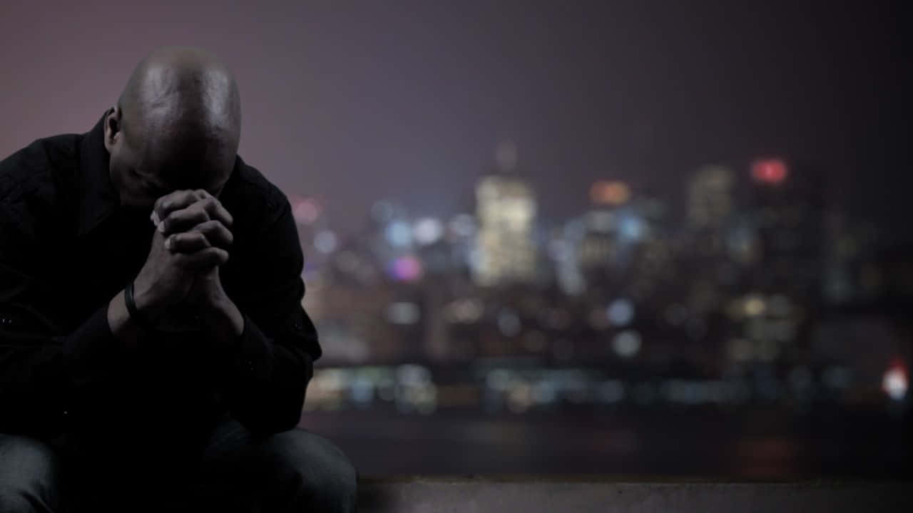 Praying 桌布 壁紙 — 一名男子在夜晚模糊的城市天際線前跪下祈禱，遠處有閃爍的燈光。