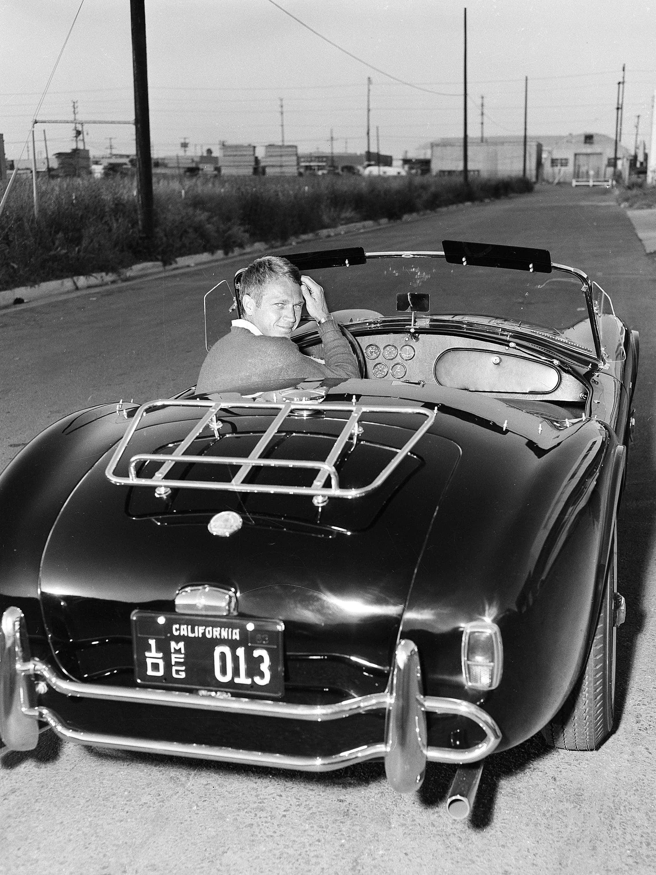 Black And White Vintage Car 桌布 壁紙 — 一名男子在復古黑色開篷車上微笑，同時在明亮的日光下於安靜的路邊整理頭髮。
