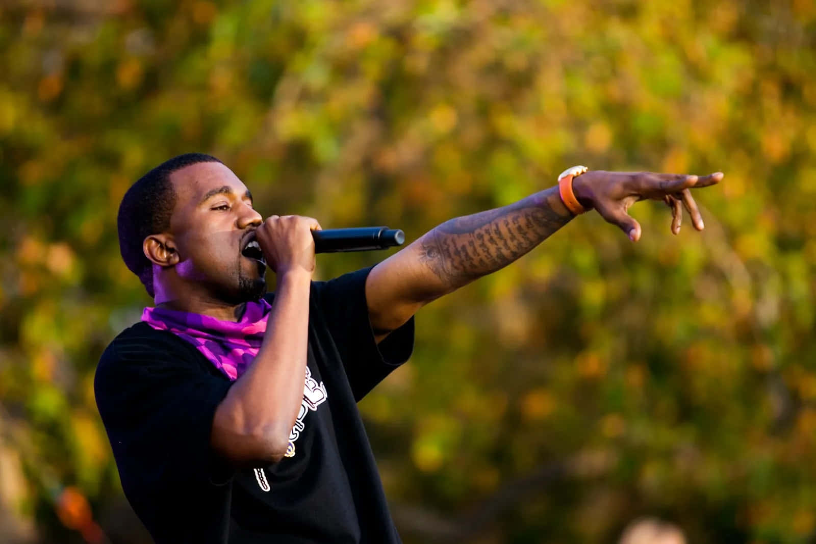 Kanye 桌布 壁紙 — 一名男子在戶外熱情地對著麥克風唱歌，身後秋天的樹葉在溫暖的金色光芒中閃耀。