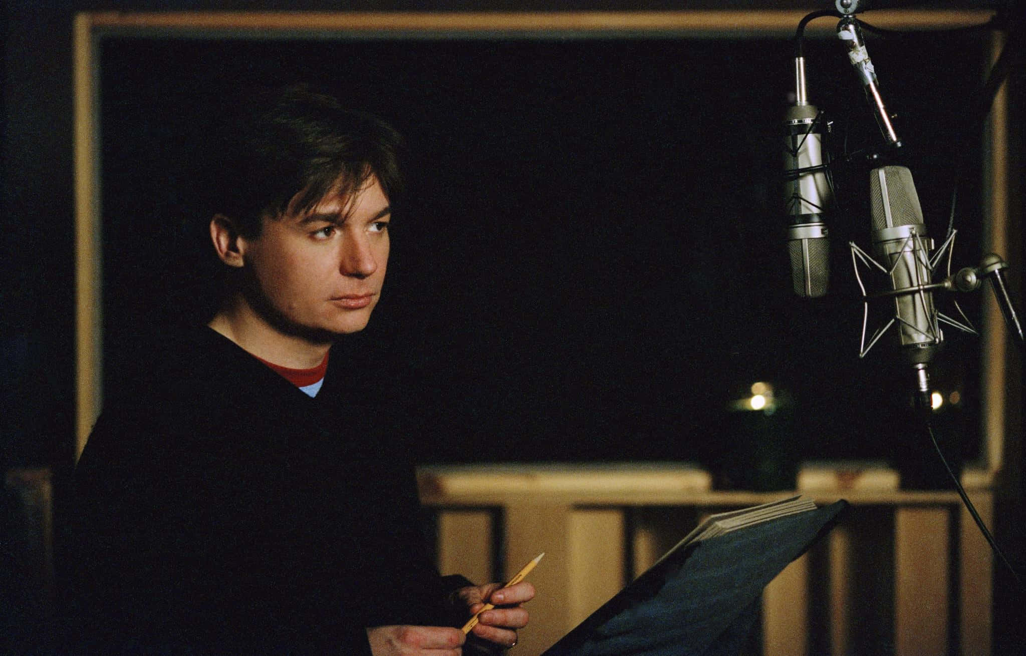 Mike Myers 桌布 壁紙 — 一名男子在昏暗的錄音室中，手持劇本和鉛筆，站在兩個專業麥克風前。