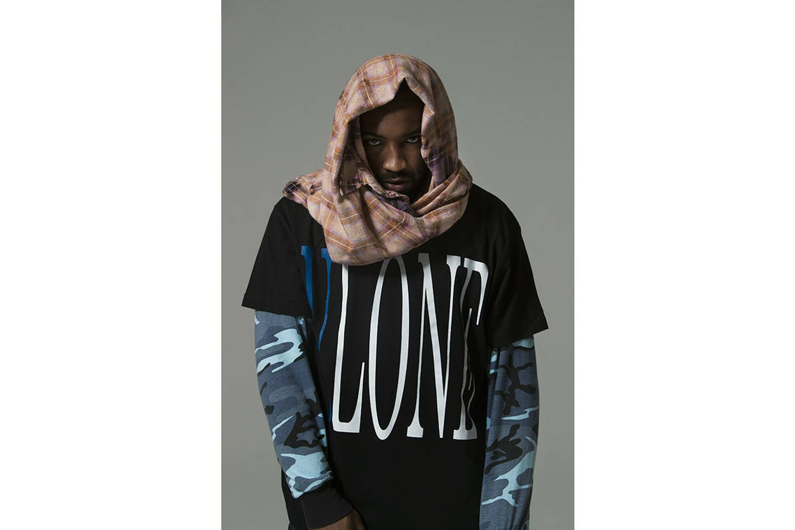 A$AP Bari x VLONE 壁紙 — 一名男子在灰色中性背景前，將格紋圍巾當作兜帽，穿在帶有粗體白色字樣和迷彩袖的黑色T恤外面。