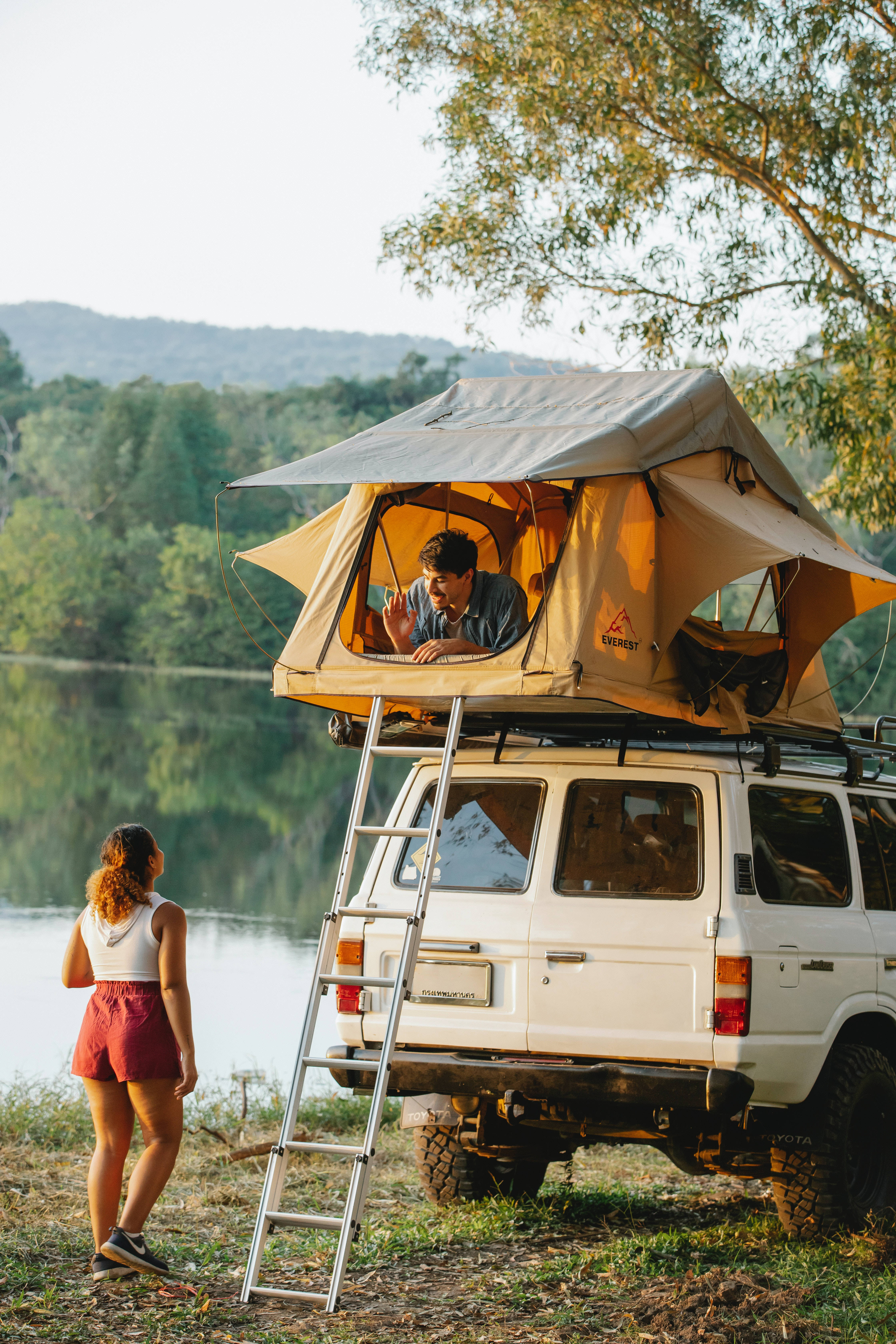 Camping 桌布 壁紙 — 一名男子在白色SUV的車頂帳篷內放鬆，而一名女子則站在下方被樹木環繞的平靜湖邊。