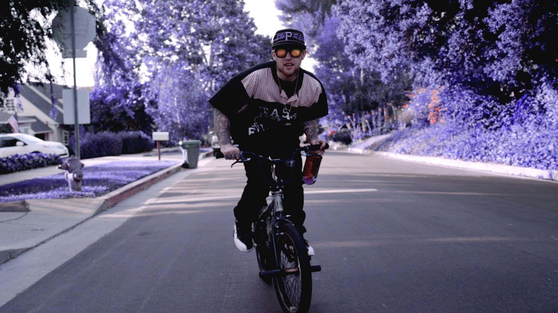 Mac Miller 桌布 壁紙 — 一名男子在超現實的紫色天空下騎單車沿著郊區街道行駛，樹木以單色調發光。