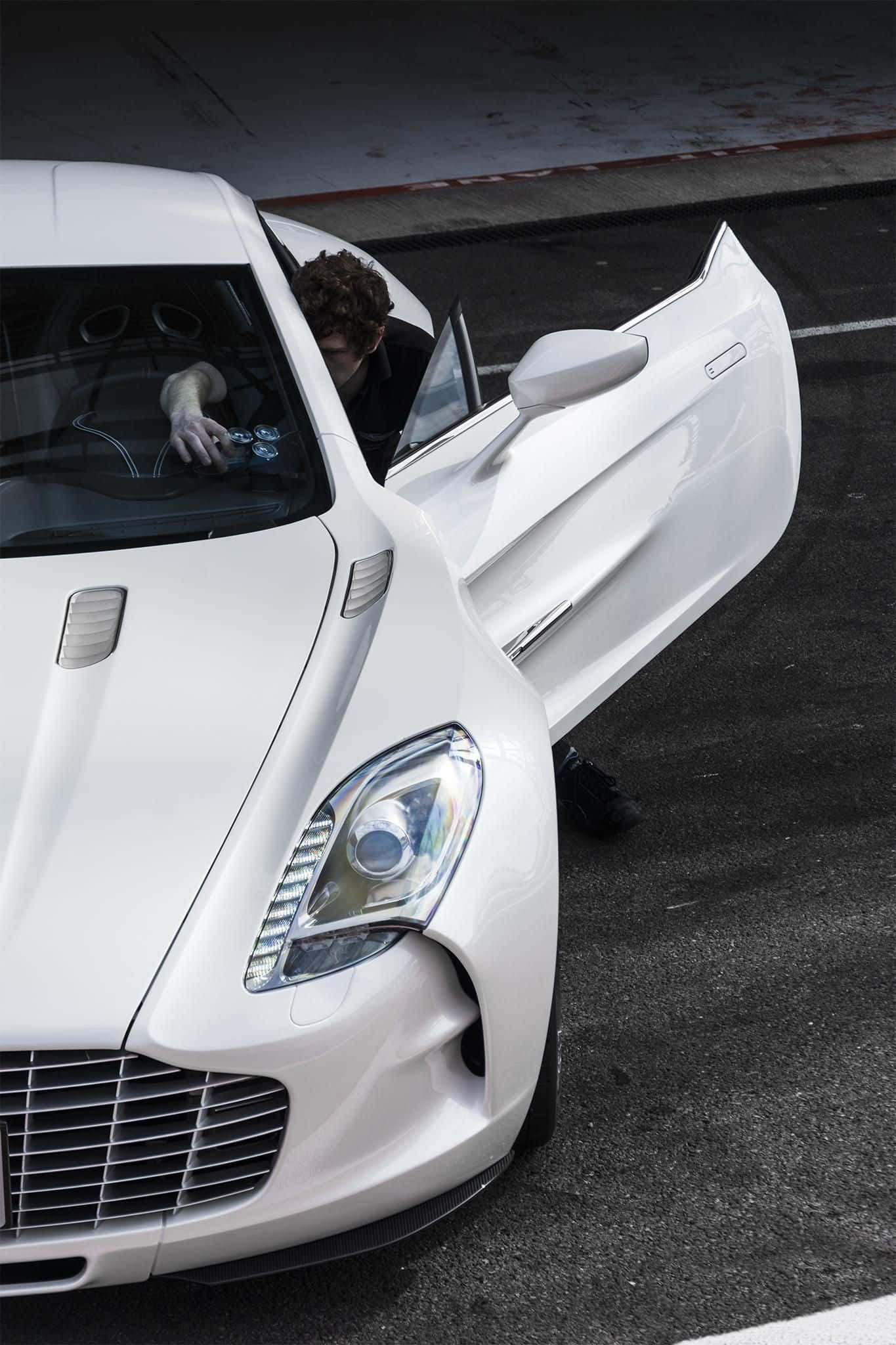 Aston Martin One-77 桌布 壁紙 — 一名男子坐在白色跑車內，鷗翼門在深色瀝青路面上打開。