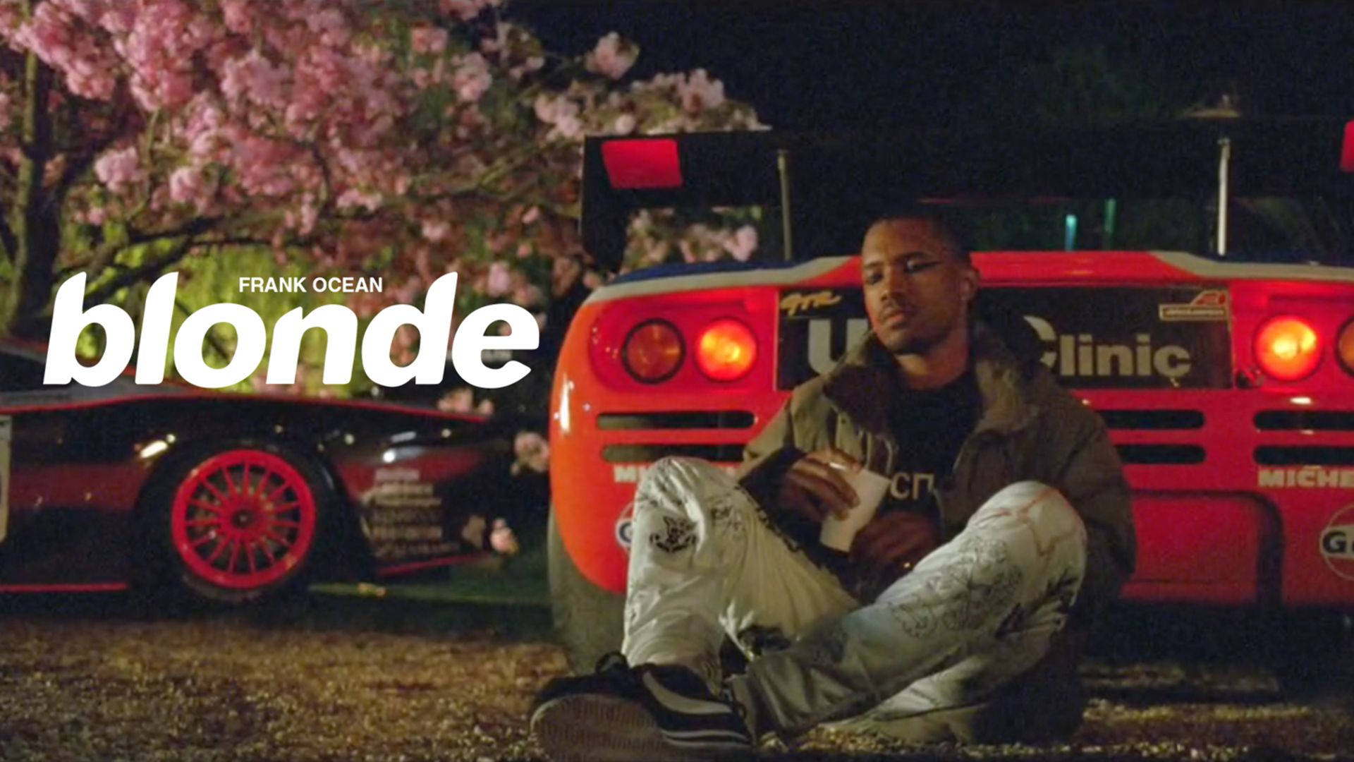 Frank Ocean《Nikes》MV 壁紙 — 一名男子晚上坐在粉紅色櫻花下的紅色賽車旁邊的地上，尾燈閃閃發光。