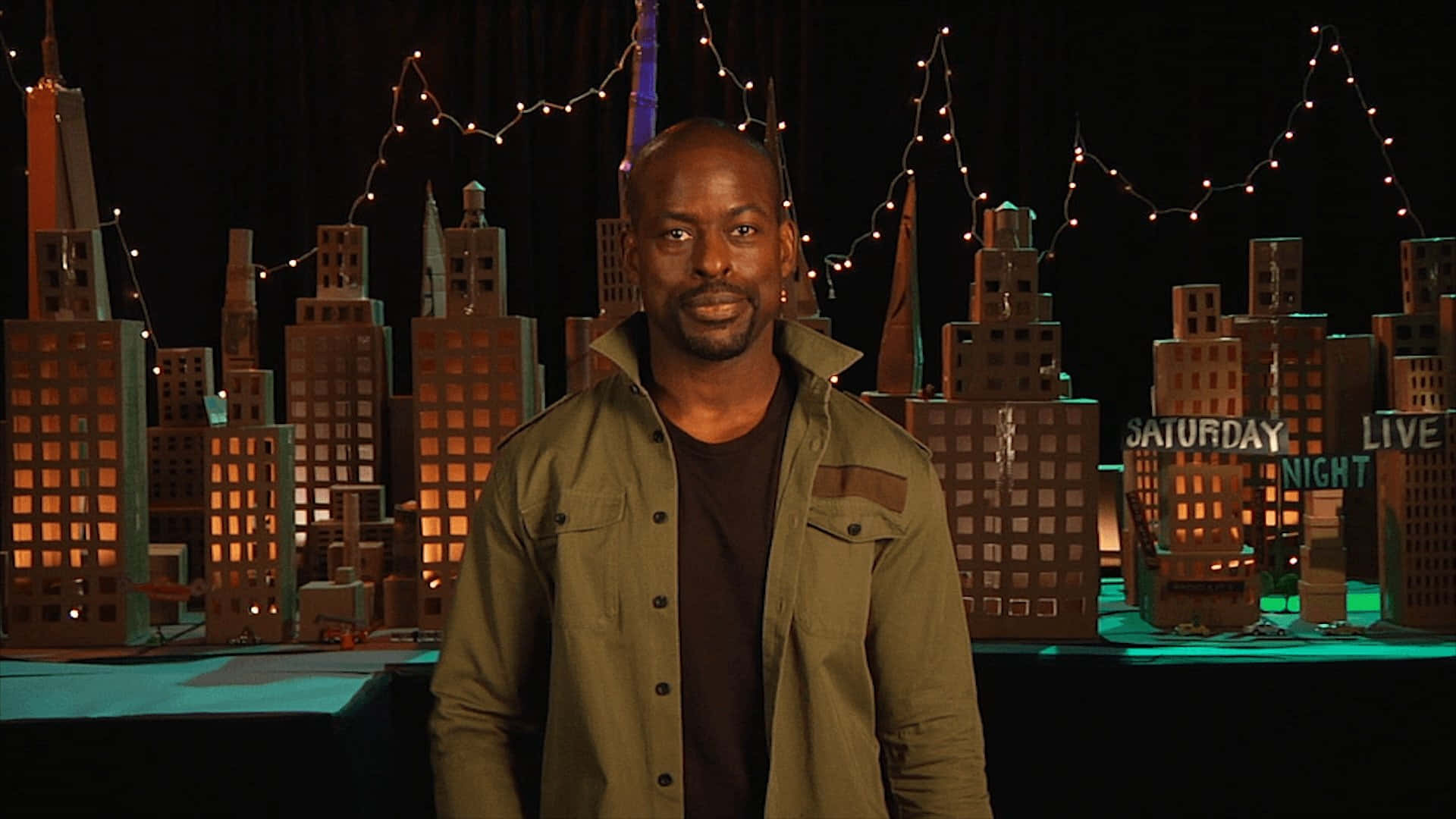 Sterling K Brown 桌布 壁紙 — 一名男子站在一個以溫暖串燈和發光窗戶照亮的微型城市背景前。