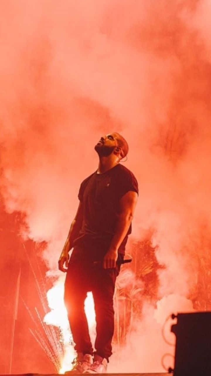 Drake 桌布 壁紙 — 一名男子站在舞台上，被熾熱的橙色煙霧和火花包圍，進行一場高能量的音樂會表演。