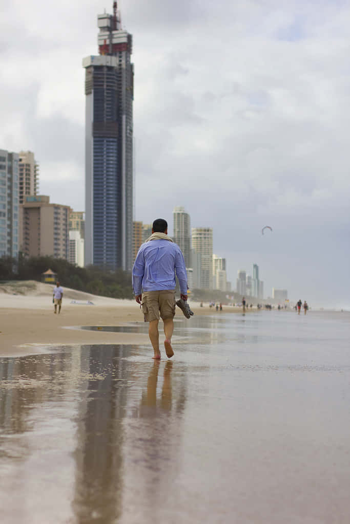 Beach Walk 桌布 壁紙 — 一名男子赤腳走在濕沙灘上，背景是陰天下一座正在建造的摩天大樓。