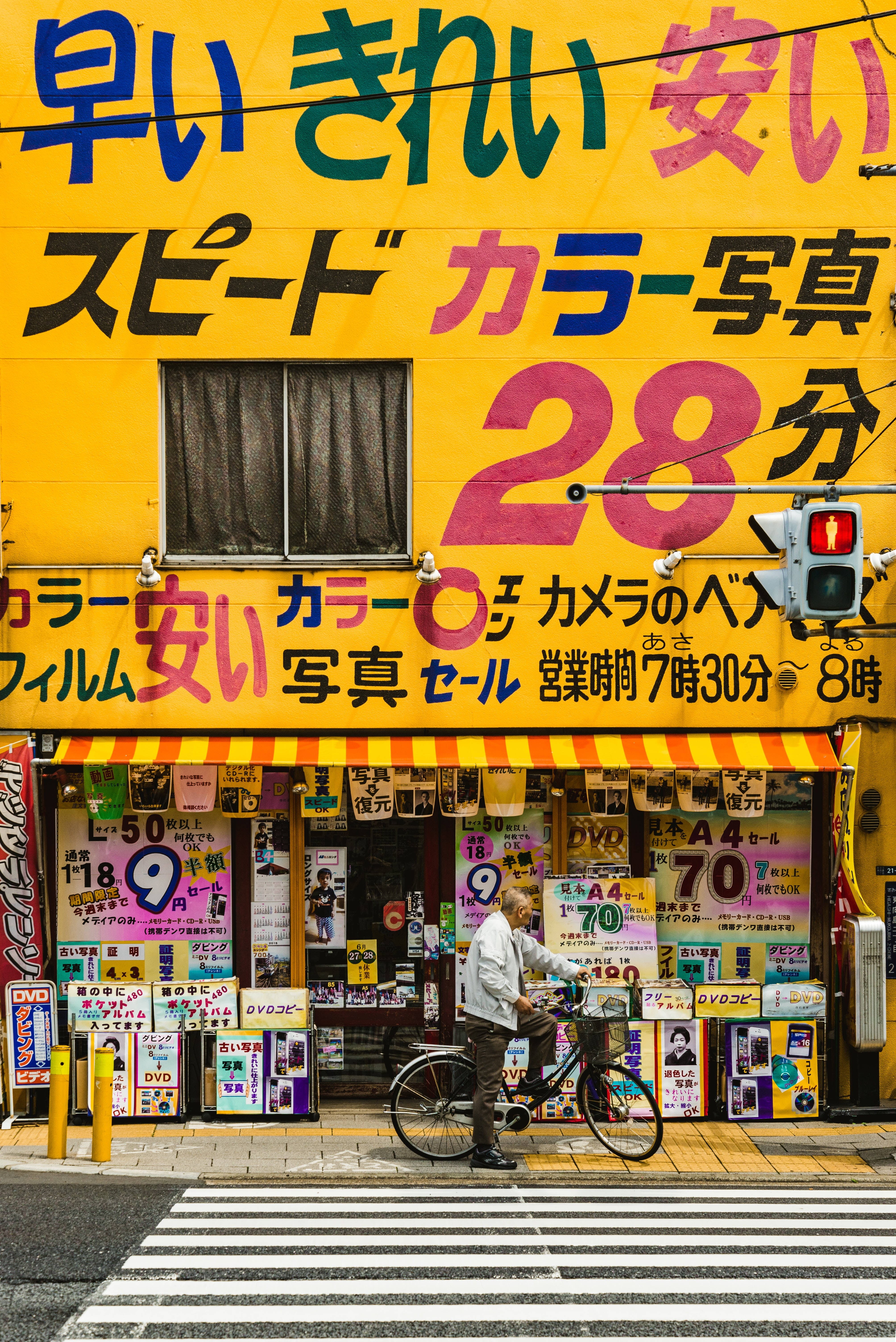 Store 桌布 壁紙 — 一名男子踩單車經過一間充滿活力的黃色日本相片店，店舖有鮮豔奪目的彩色招牌和一個紅色交通燈。