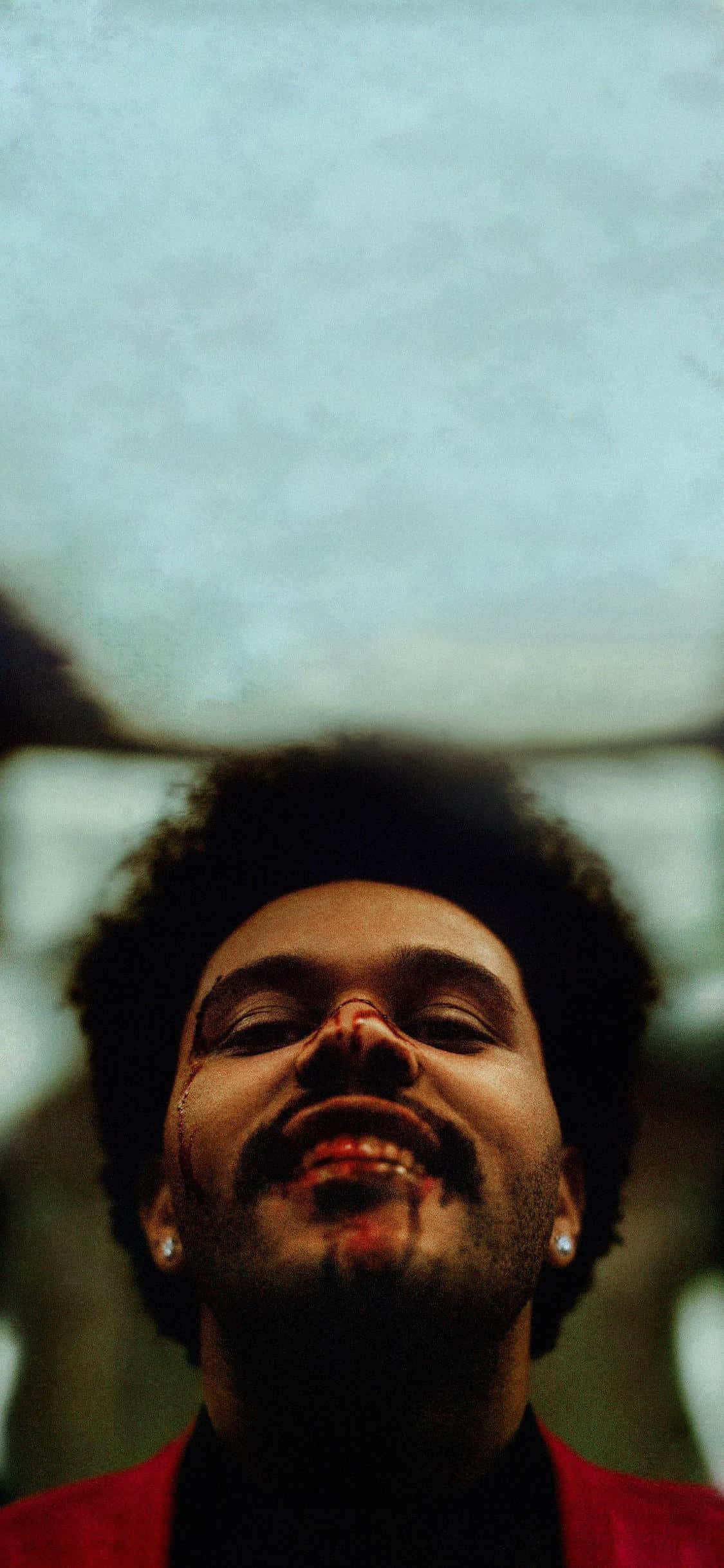 The Weeknd 桌布 壁紙 — 一名男子閉上眼睛，露出愉快的笑容，身穿紅色外套，背景模糊的特寫肖像。