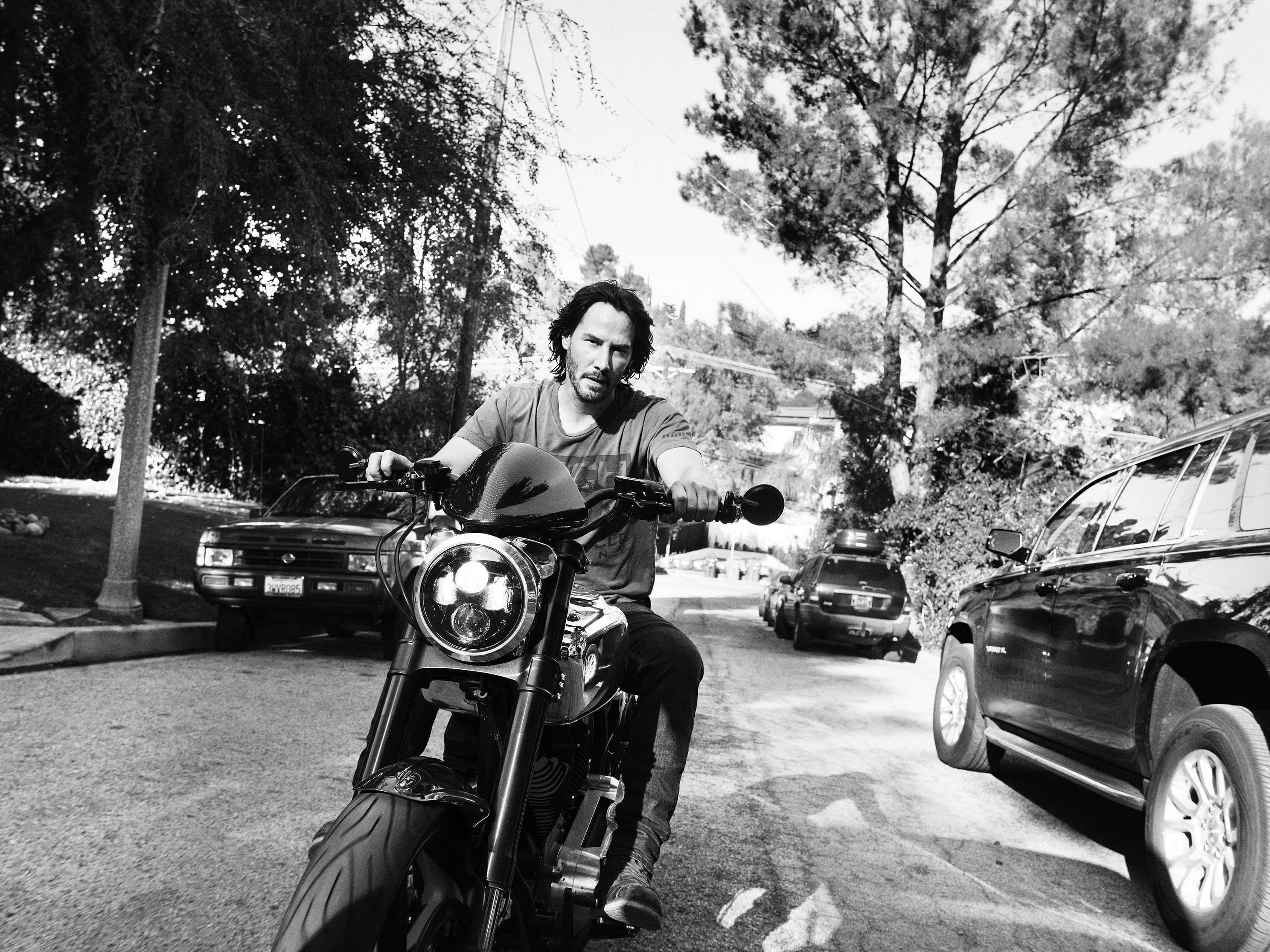 Keanu Reeves 桌布 壁紙 — 一名男子騎著電單車，沿著兩旁有樹木的住宅街道行駛，黑白畫面，兩邊停有汽車。