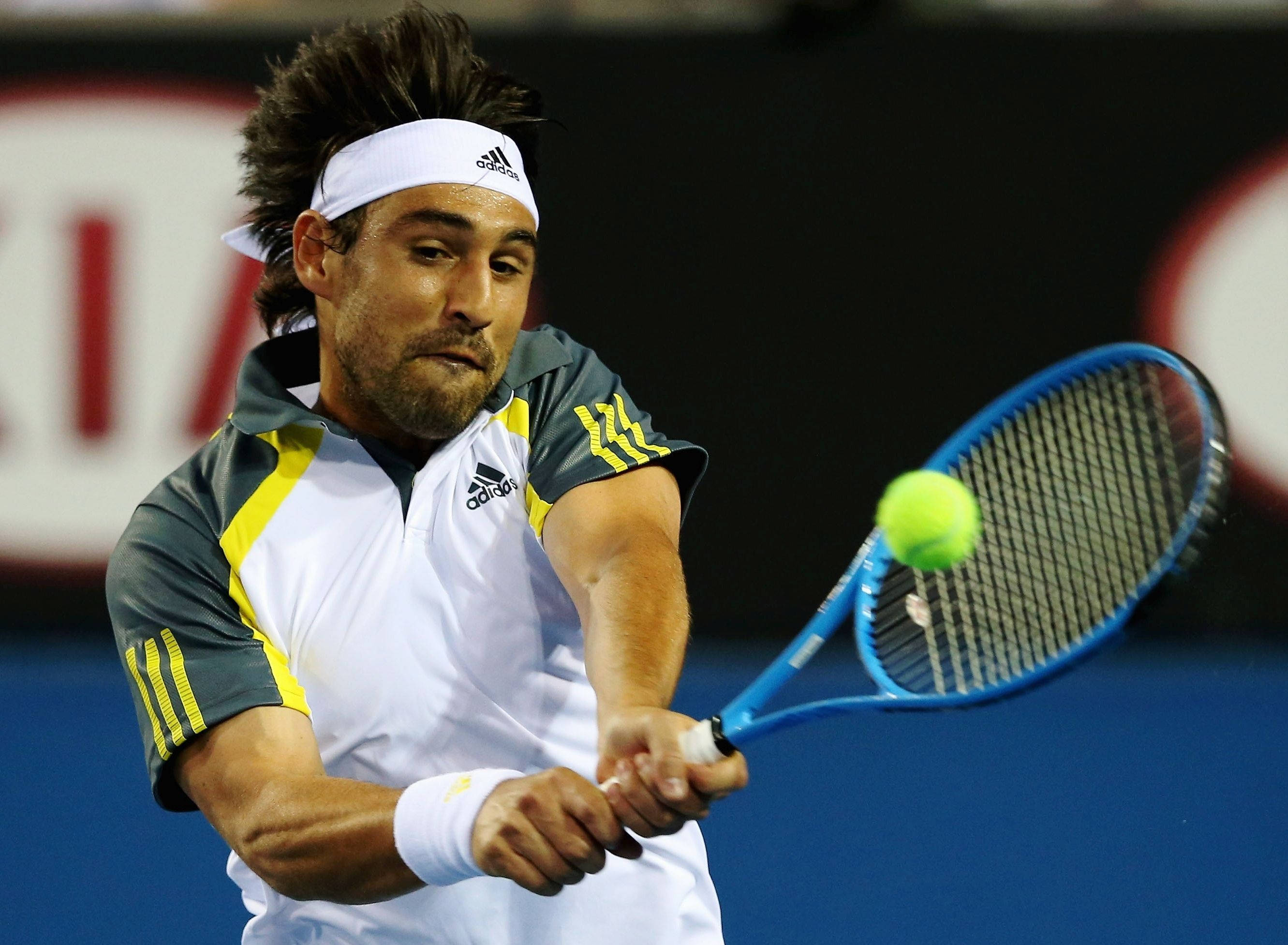 Marcos Baghdatis Hitting Ball 壁紙 — 一名男性網球運動員在揮拍中擊中一個亮黃色的球，使用藍色球拍，背景為深色。