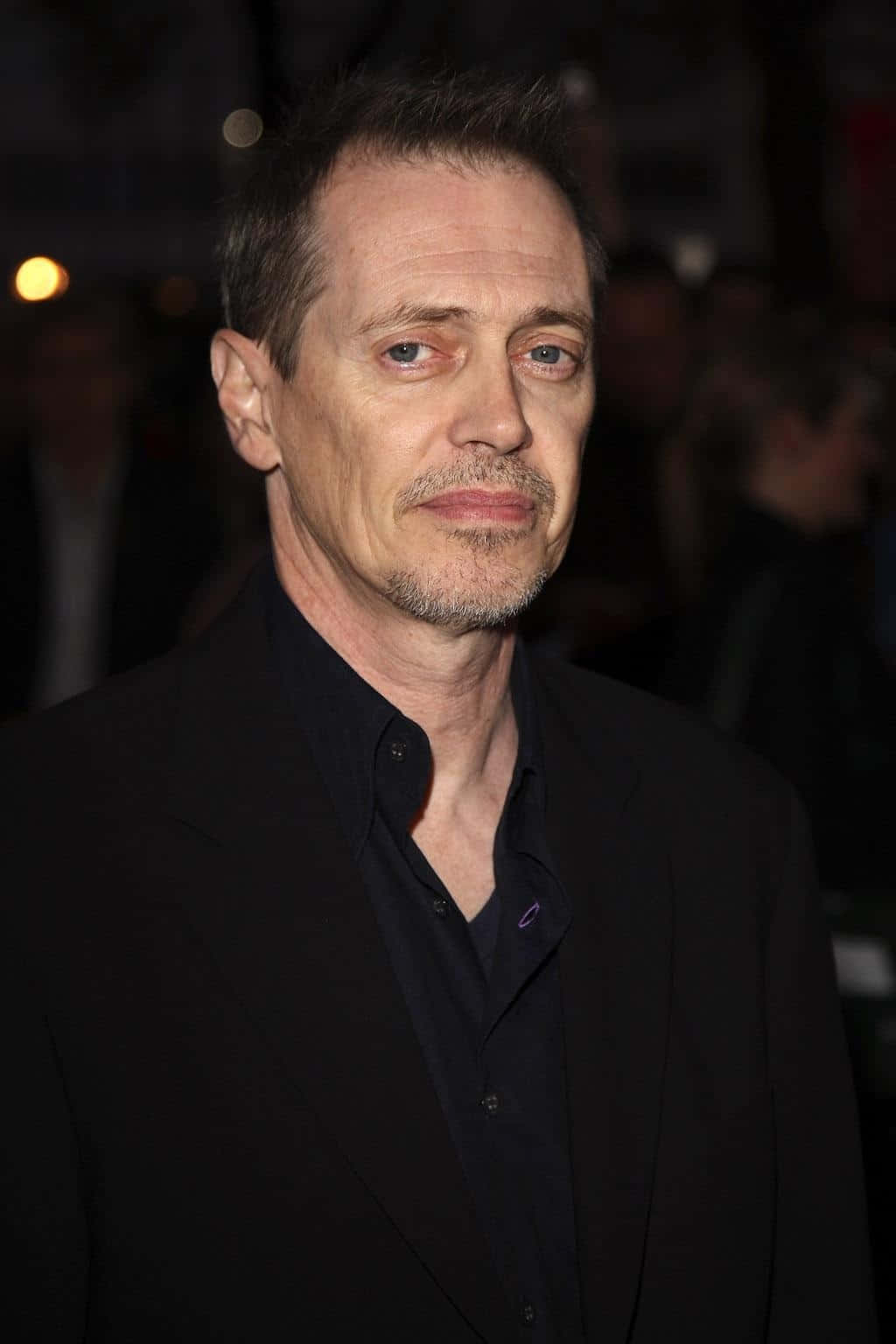 steve buscemi 桌布 壁紙 — 一名留著灰色鬍渣、擁有深邃藍眼睛的男子特寫肖像，身穿黑色西裝和襯衫，背景是模糊的夜色。