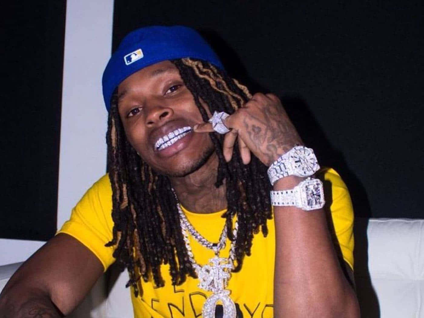 King Von 桌布 壁紙 — 一名留著辮子頭、佩戴鑽石首飾的男子在昏暗的房間裡微笑著觸摸自己的牙齒。