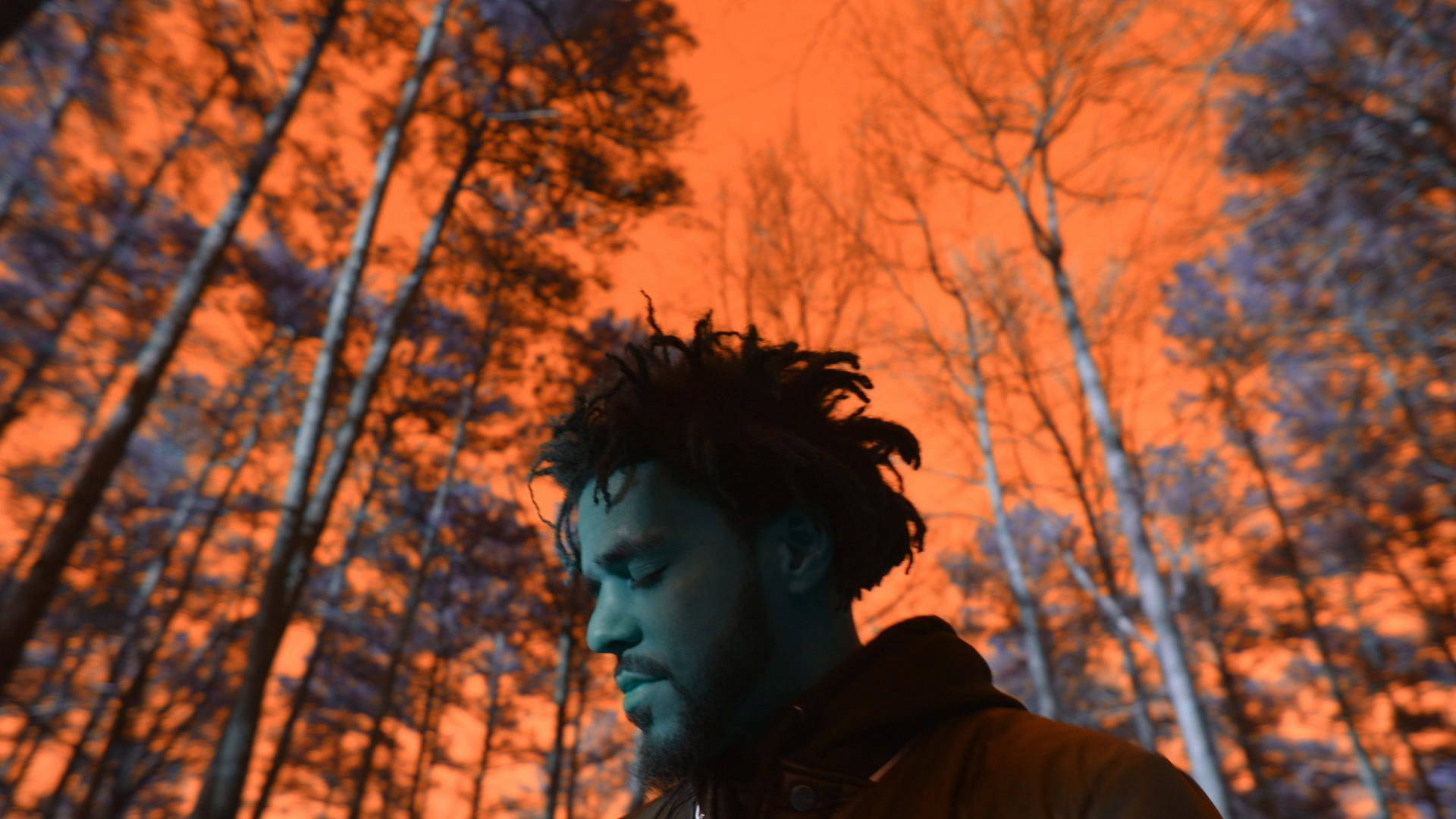 J Cole《4 Your Eyez Only》 壁紙 — 一名留著辮子頭的男人在森林中，在強烈的橙色天空下向下凝視。
