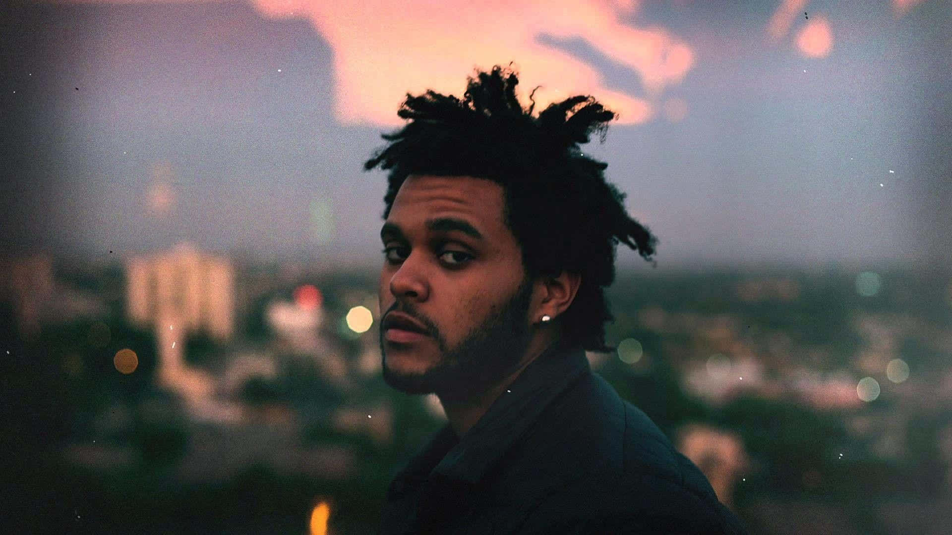 The Weeknd After Hours 桌布 壁紙 — 一名留著辮子頭的男子，專注地凝視著在夕陽色調和散景燈光下閃爍的模糊城市天際線。