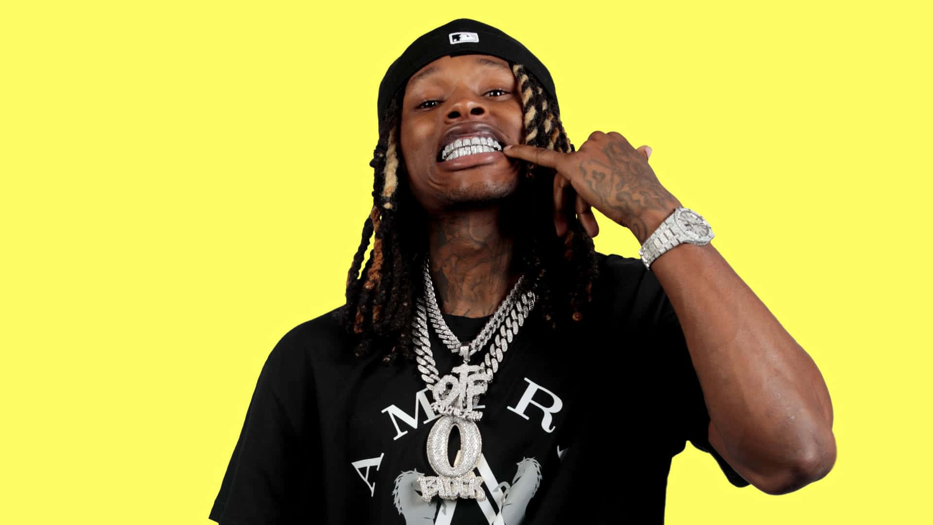 King Von 桌布 壁紙 — 一名留著辮子頭髮、戴著金鏈的男子在明亮的黃色背景前自信地擺姿勢。