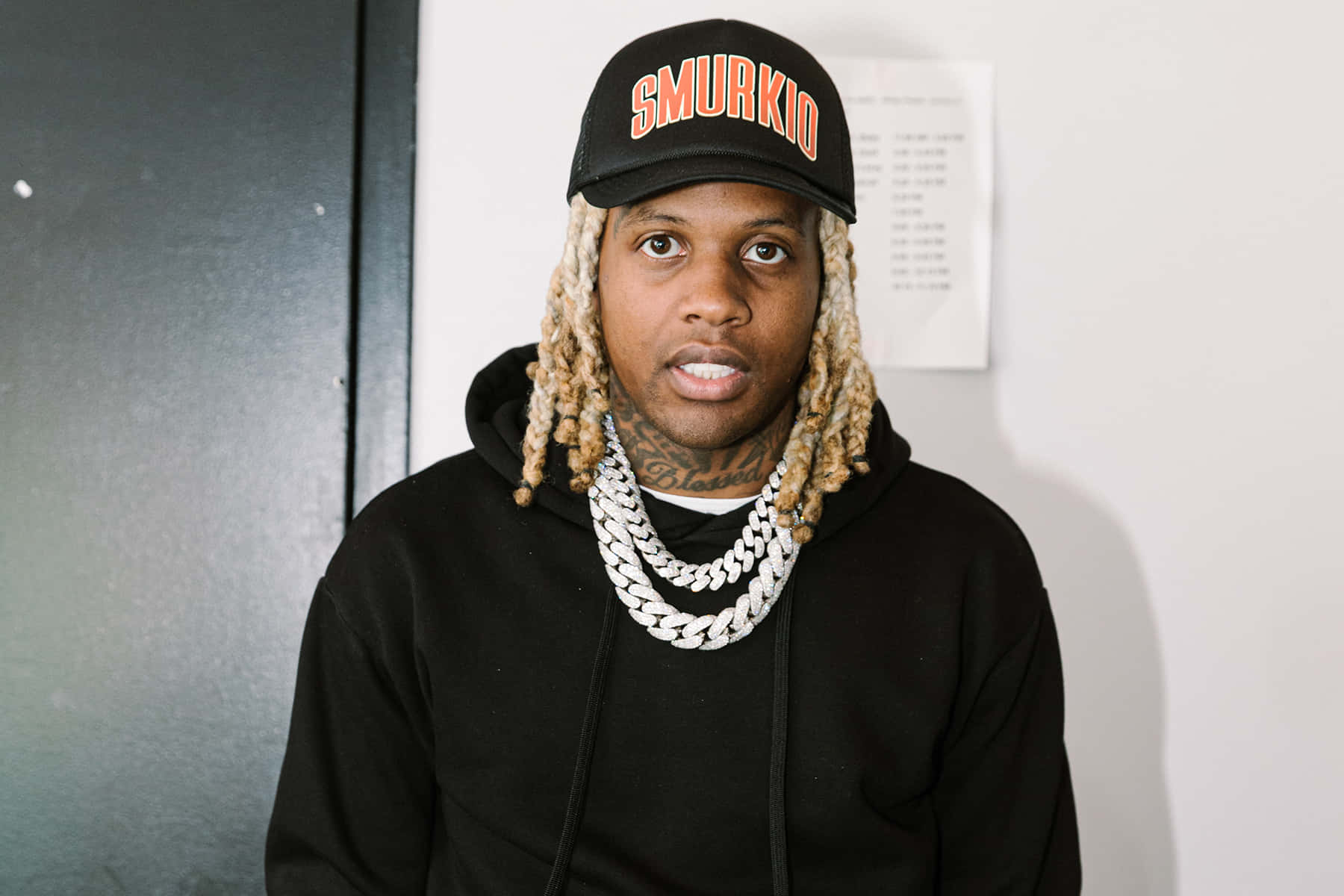 lil durk 桌布 壁紙 — 一名留著金色辮子、戴著黑色「SMURKIO」帽子的男子，身穿黑色連帽衫和粗銀鏈，直視鏡頭。