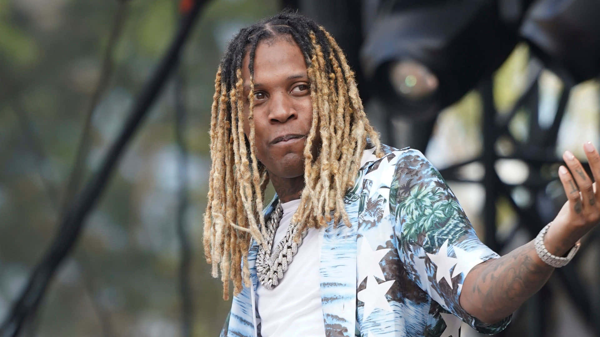 lil durk 桌布 壁紙 — 一名留著金色長辮子的男子，穿著熱帶印花襯衫和粗鏈條項鍊，做著手勢。