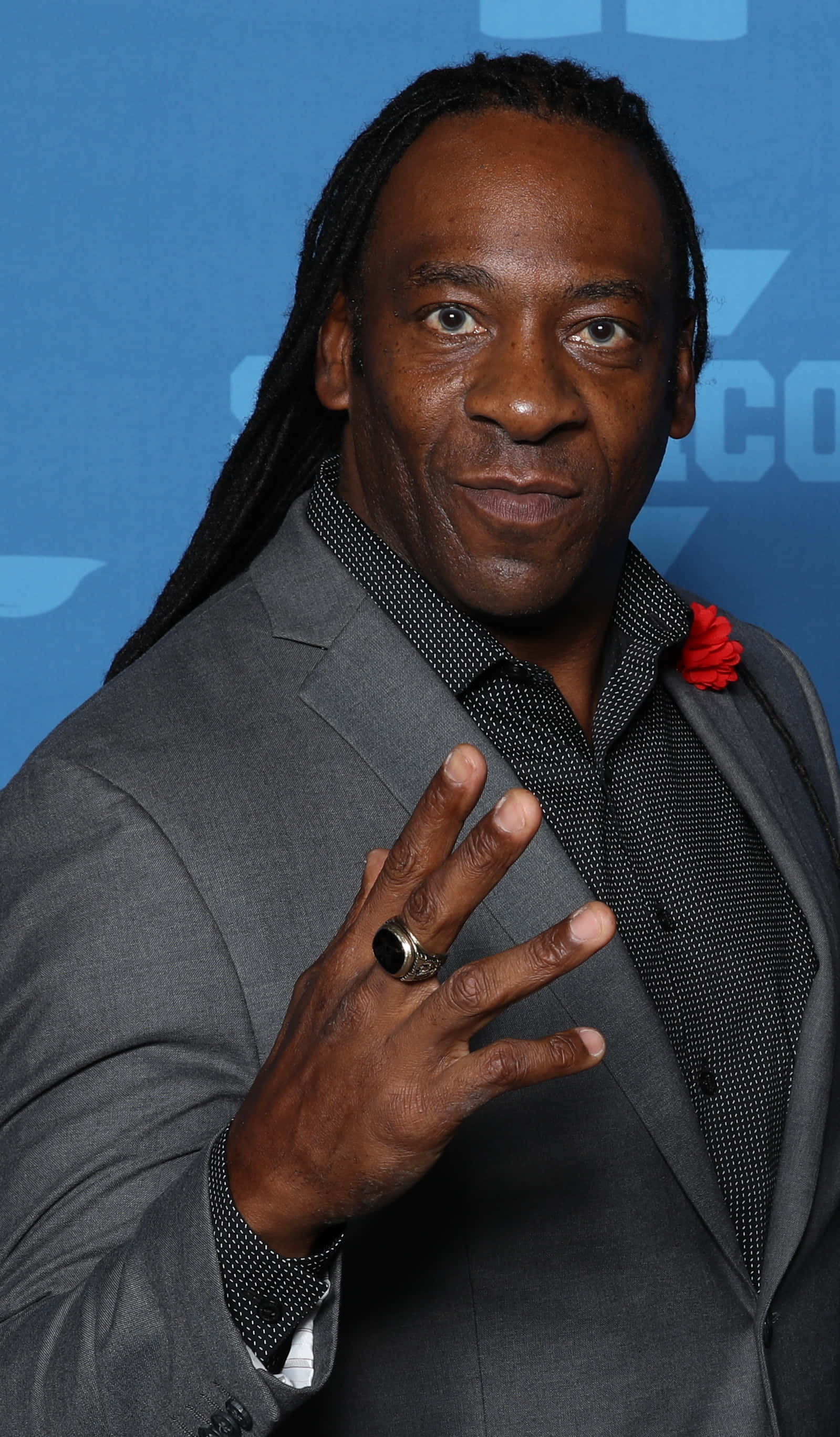 "Booker T, The Legendary WWE Wrestler, in a Dynamic Pose (2018)" 壁紙 — 一名留著長辮子、身穿灰色西裝的男子在藍色背景前微笑並做出手勢。
