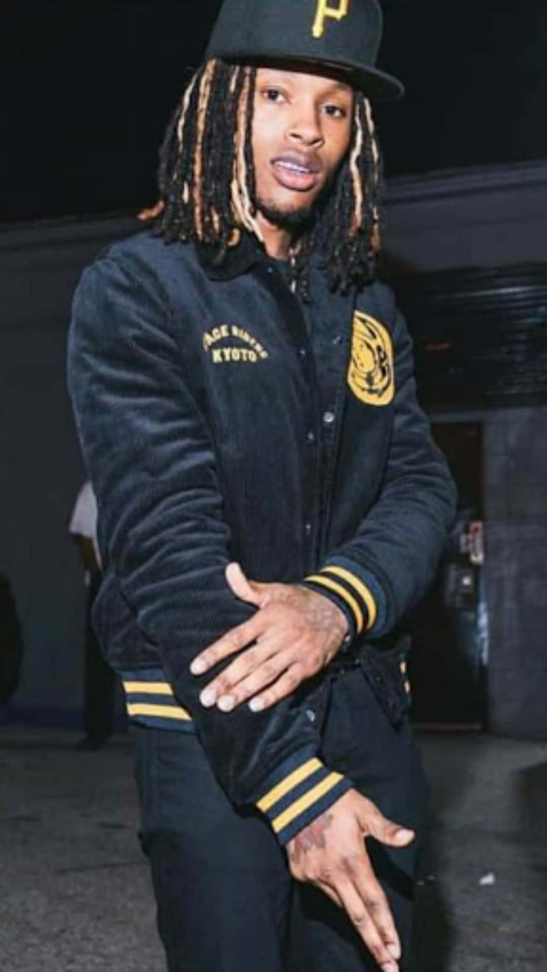 King Von 桌布 壁紙 — 一名留著長辮子的男人，在夜間城市燈光下穿著一件帶有黃色條紋和補丁的黑色天鵝絨校隊夾克。