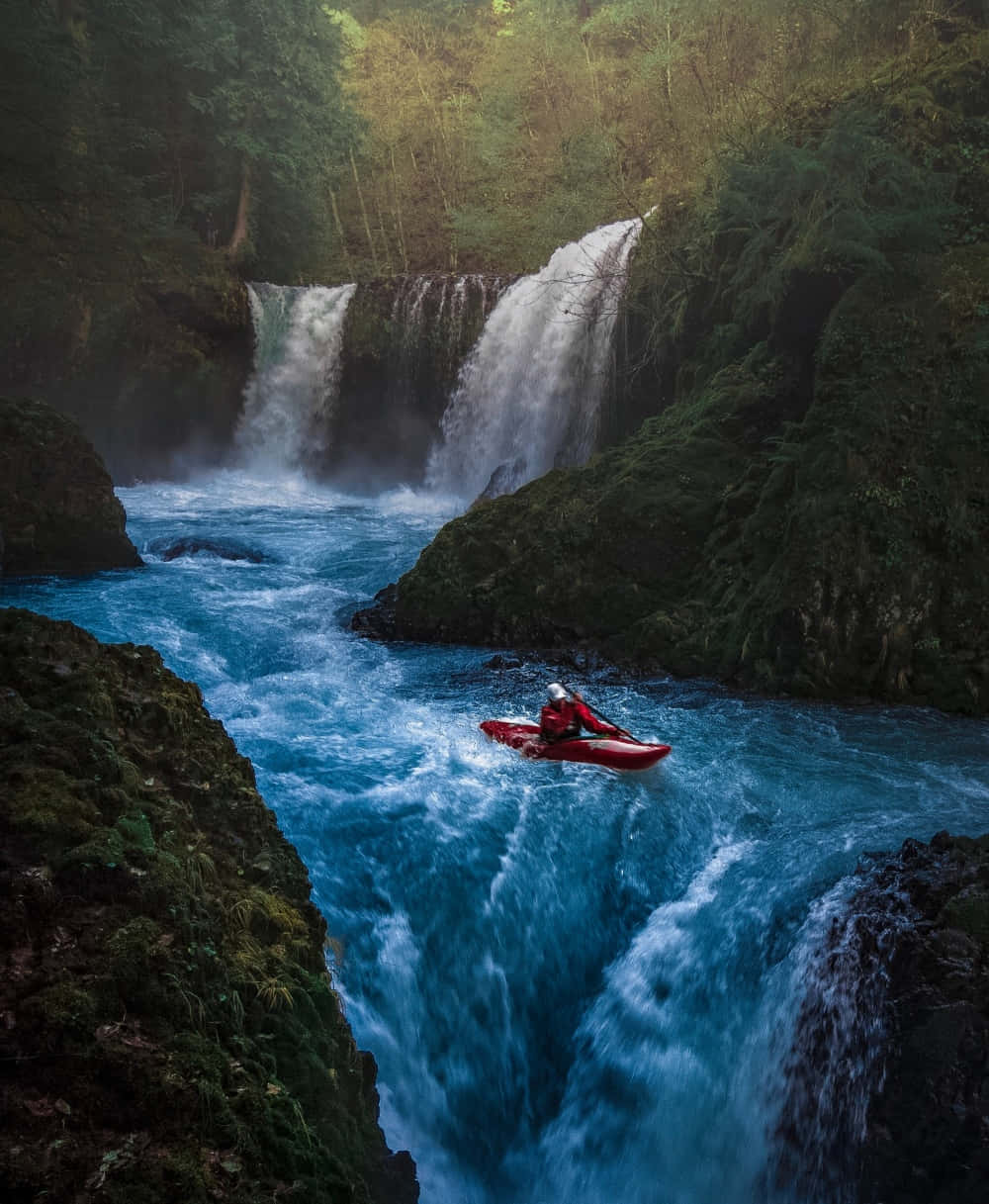 adventure 桌布 壁紙 — 一名皮划艇運動員在紅色船上，在翠綠森林環繞的瀑布下，於湍急的藍色水域中航行。