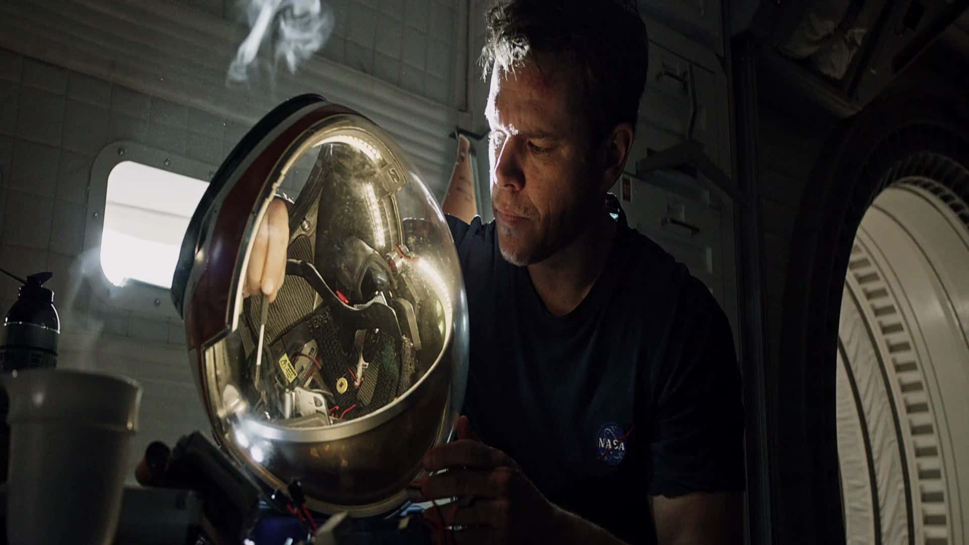 The Martian 桌布 壁紙 — 一名穿著NASA襯衫的男子在太空船艙內仔細檢查一個反光的太空人頭盔。