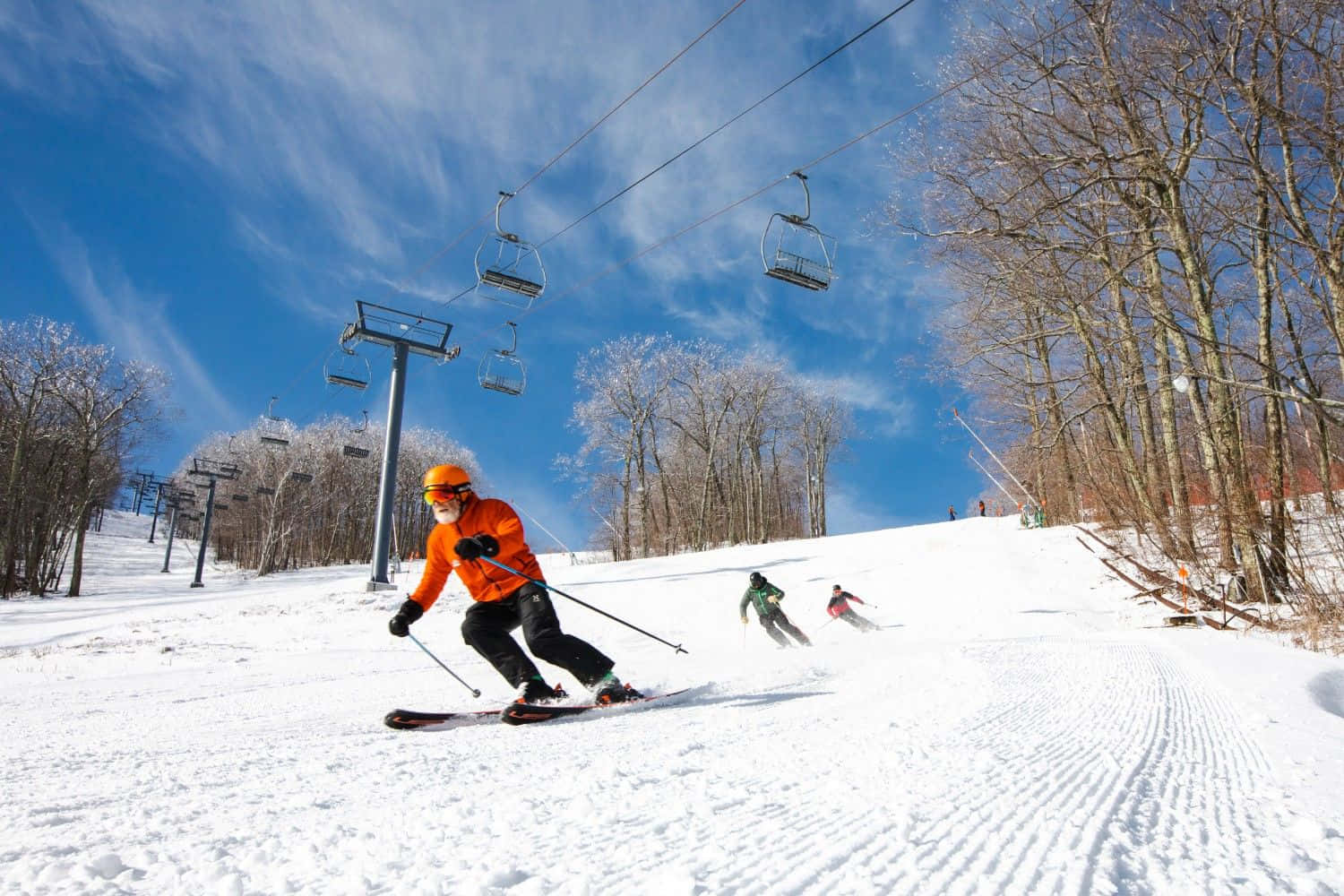 Skiing 桌布 壁紙 — 一名穿著亮橙色外套的滑雪者，在晴朗的藍天下，頭頂有纜車，沿著雪坡滑下。