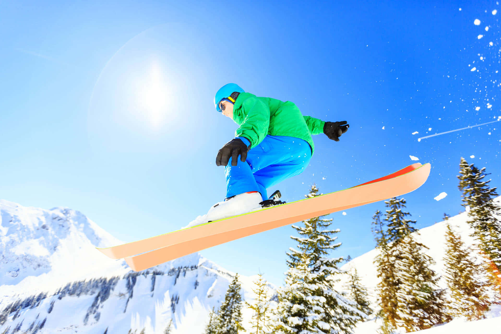 Skiing 桌布 壁紙 — 一名穿著亮綠色和藍色裝備的滑雪板運動員在燦爛的陽光下，飛越積雪的松樹，穿梭於清爽的冬日空氣中。