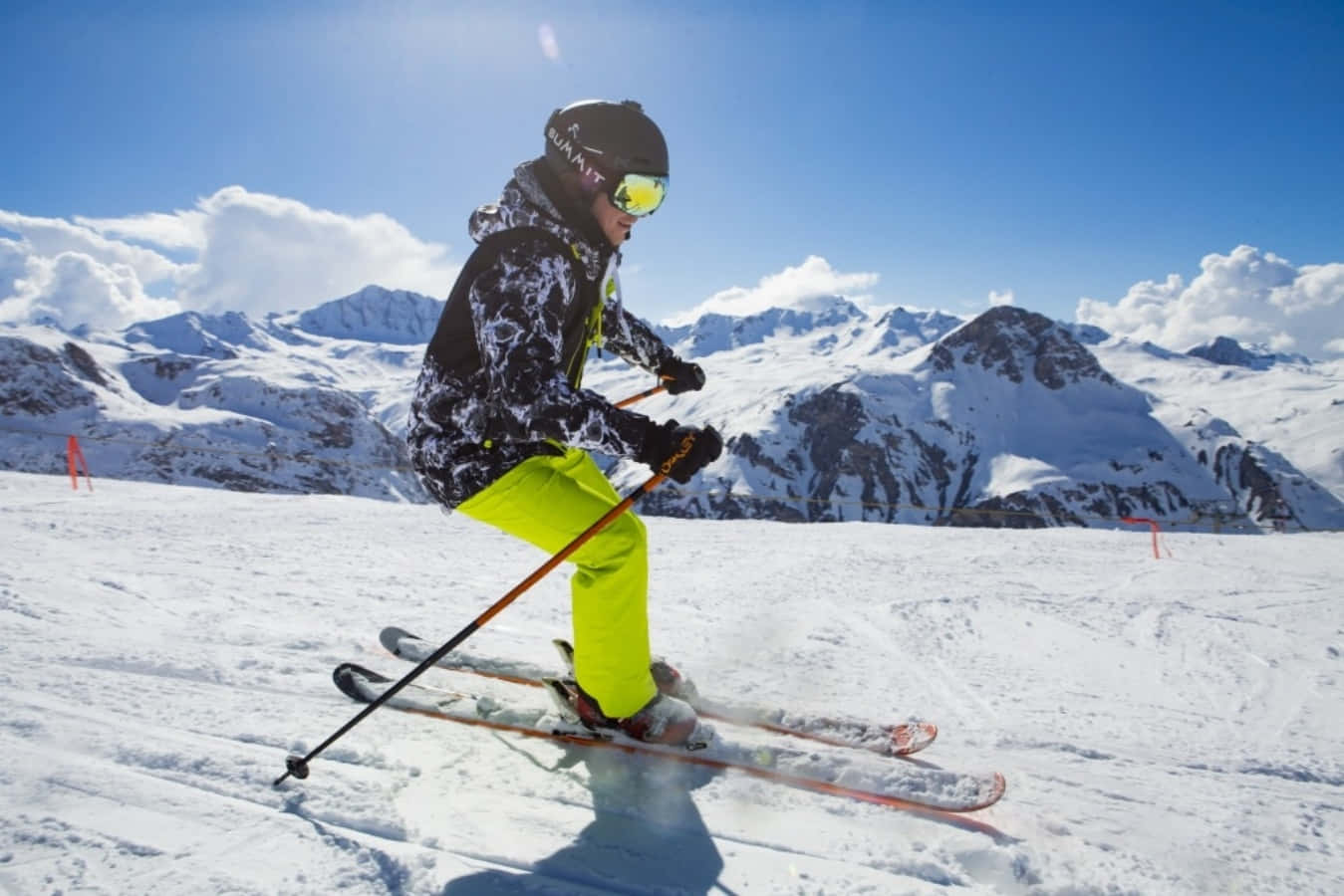 Skiing 桌布 壁紙 — 一名穿著亮黃色褲子的滑雪者，在晴朗的藍天下，背景有鋸齒狀山峰，沿著白雪覆蓋的山坡滑下。