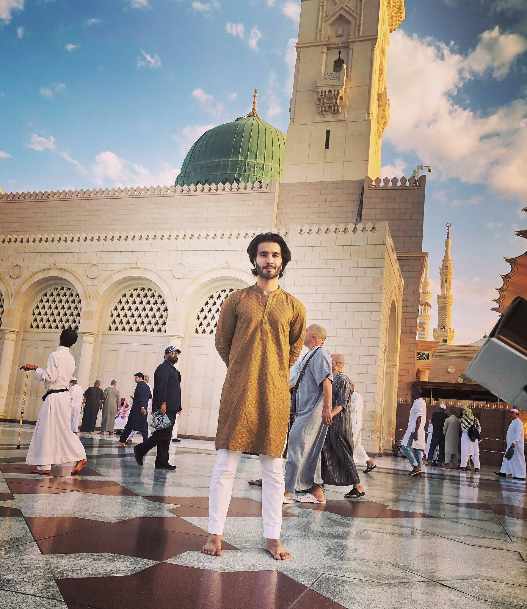 Madina Sharif 桌布 壁紙 — 一名穿著傳統服飾的年輕男子赤腳站在有圖案的庭院地上，面前是一座擁有綠色圓頂和金色宣禮塔的宏偉清真寺，背景是明亮的藍天。