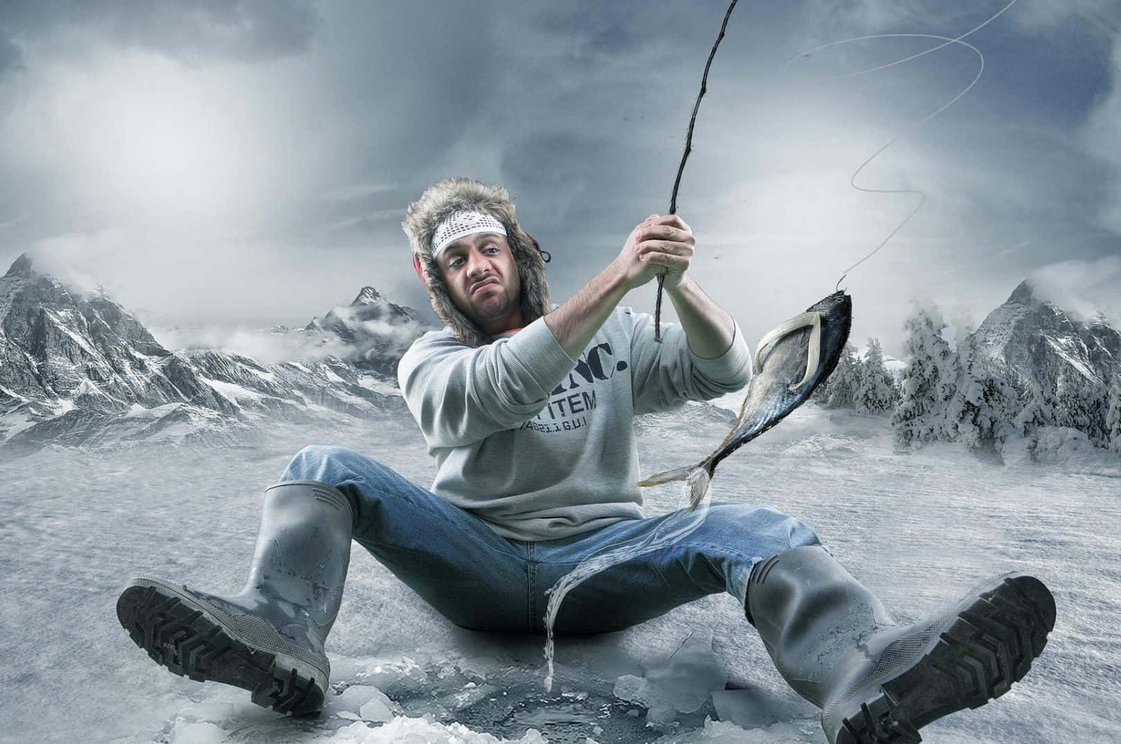 Ice Fishing 桌布 壁紙 — 一名穿著冬季裝備的男子坐在結冰的冰面上，用釣竿從洞中拉出一條大魚，背景是陰天下的雪山。