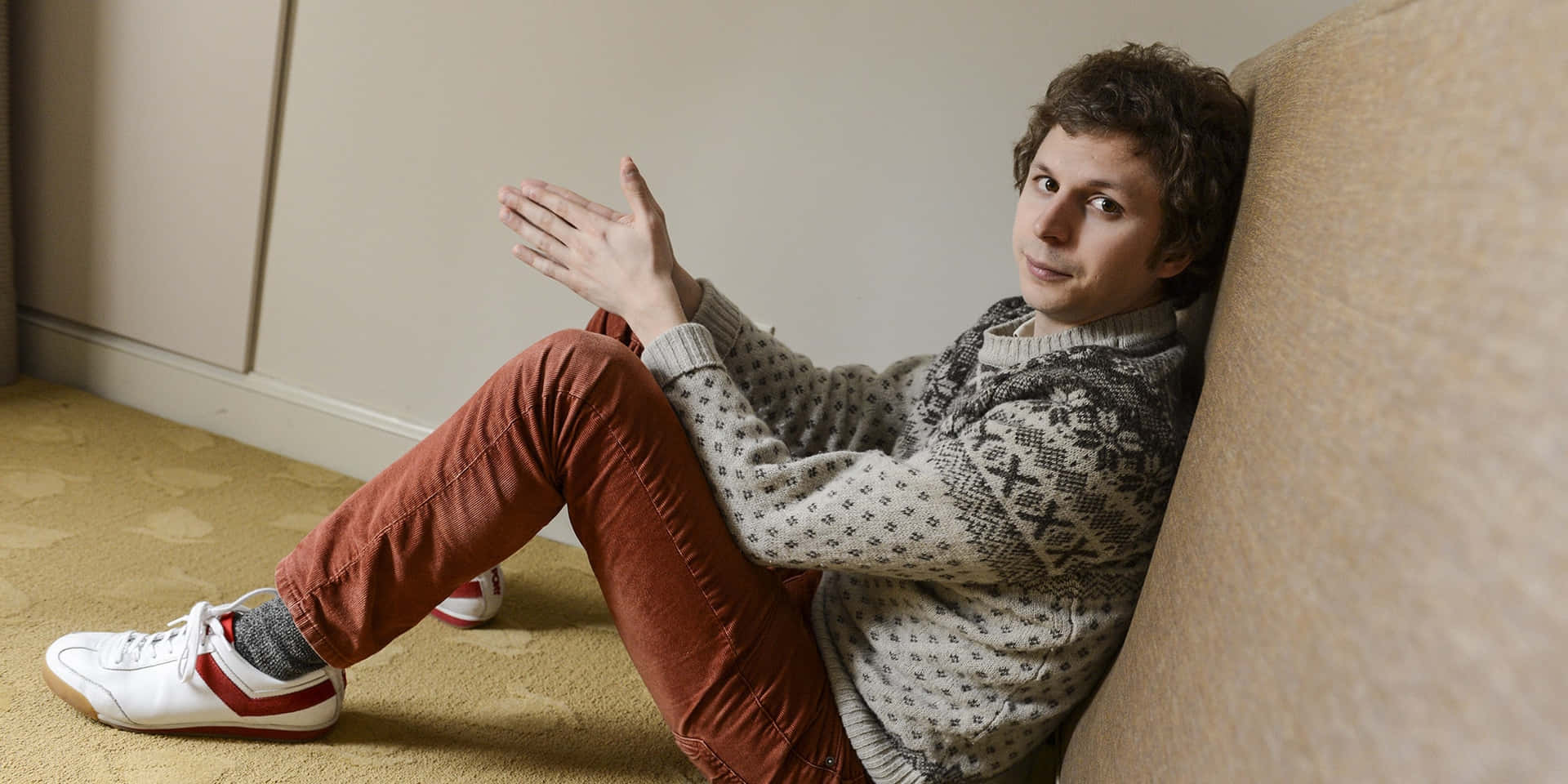 michael cera 桌布 壁紙 — 一名穿著圖案毛衣和鏽色褲子的年輕男子，隨意地坐在鋪有地毯的地板上，靠著米色沙發。