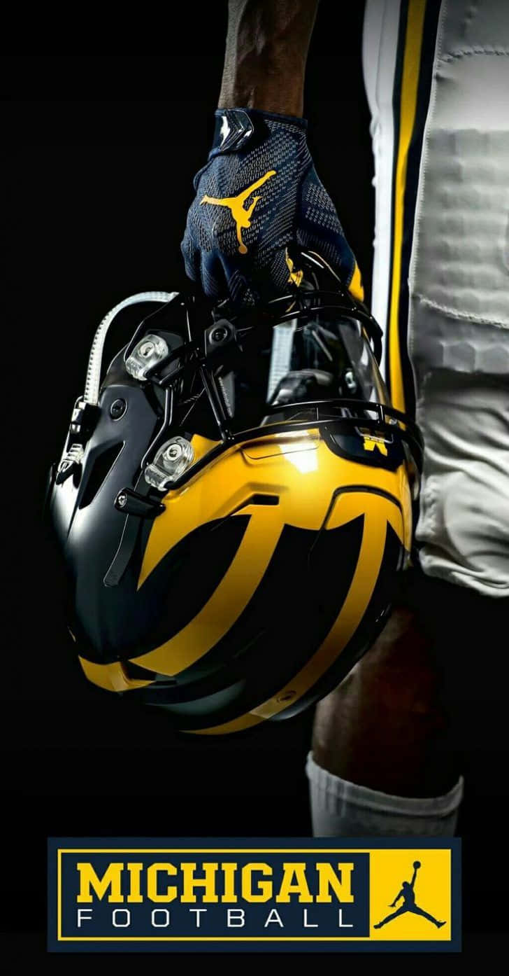 Michigan Wolverines 桌布 壁紙 — 一名穿著密歇根裝備的足球員在深色背景前手持黑黃色頭盔，背景有球隊標誌。