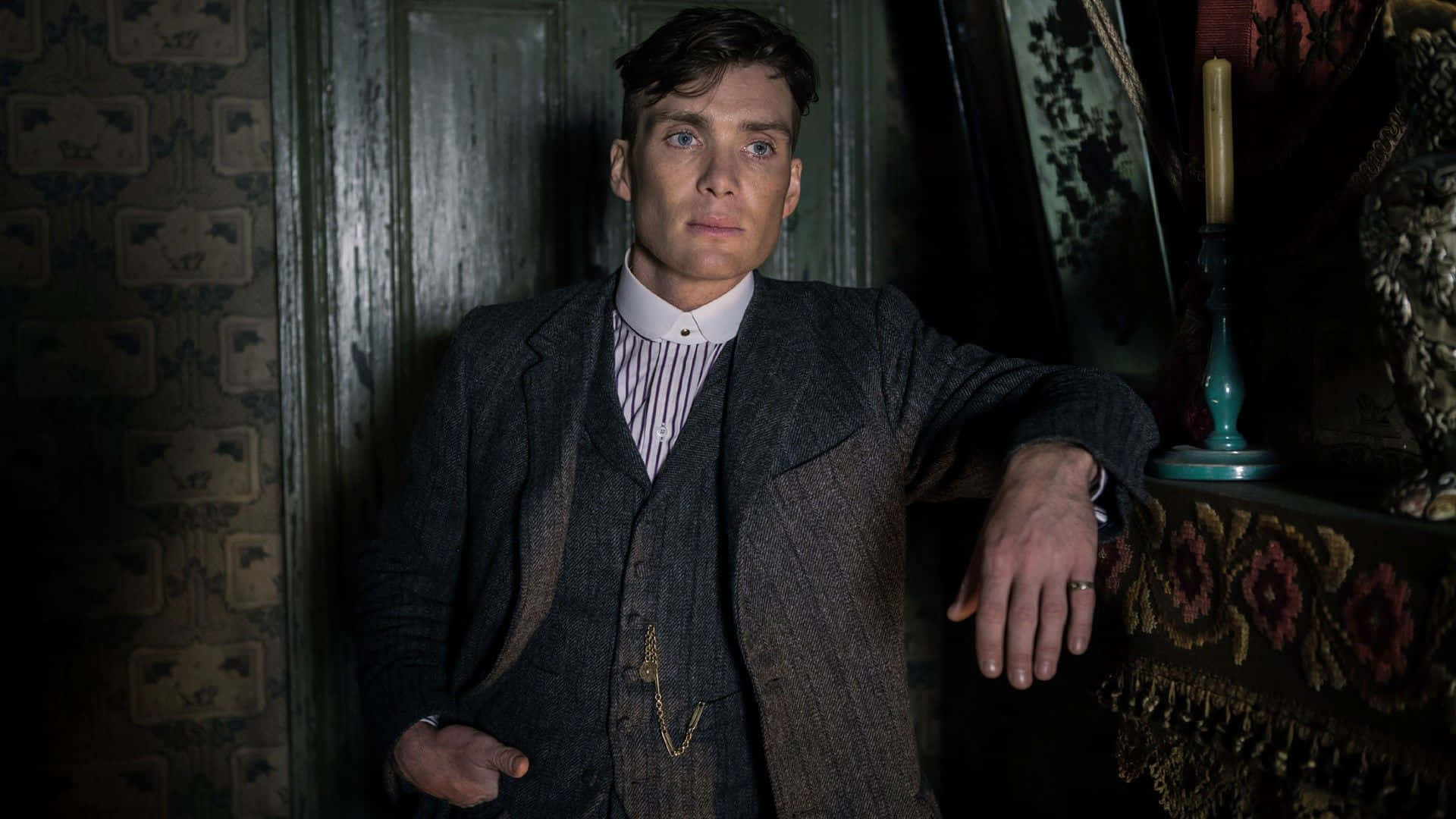 Peaky Blinders 桌布 壁紙 — 一名穿著復古西裝的男子靠在一扇深色木門上，房間昏暗華麗，舖有圖案壁紙，旁邊的桌子上點著蠟燭。