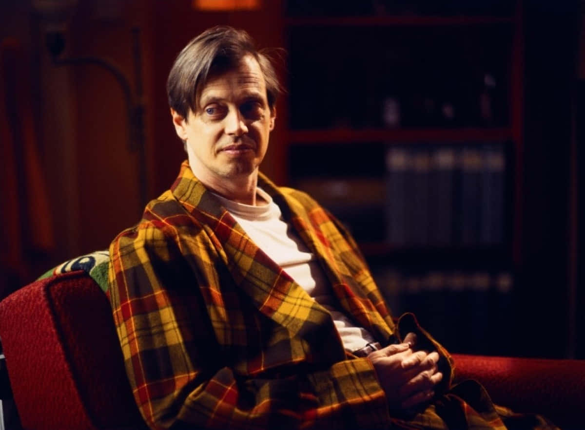 steve buscemi 桌布 壁紙 — 一名穿著格子睡袍的男子放鬆地坐在紅色扶手椅上，身後有溫暖的燈光和書架。