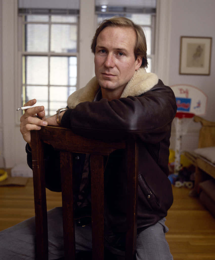 William Hurt 桌布 壁紙 — 一名穿著棕色羊羔絨外套的男子坐在木椅上，在陽光充足的房間裡拿著香煙，周圍有玩具和裱框藝術品。