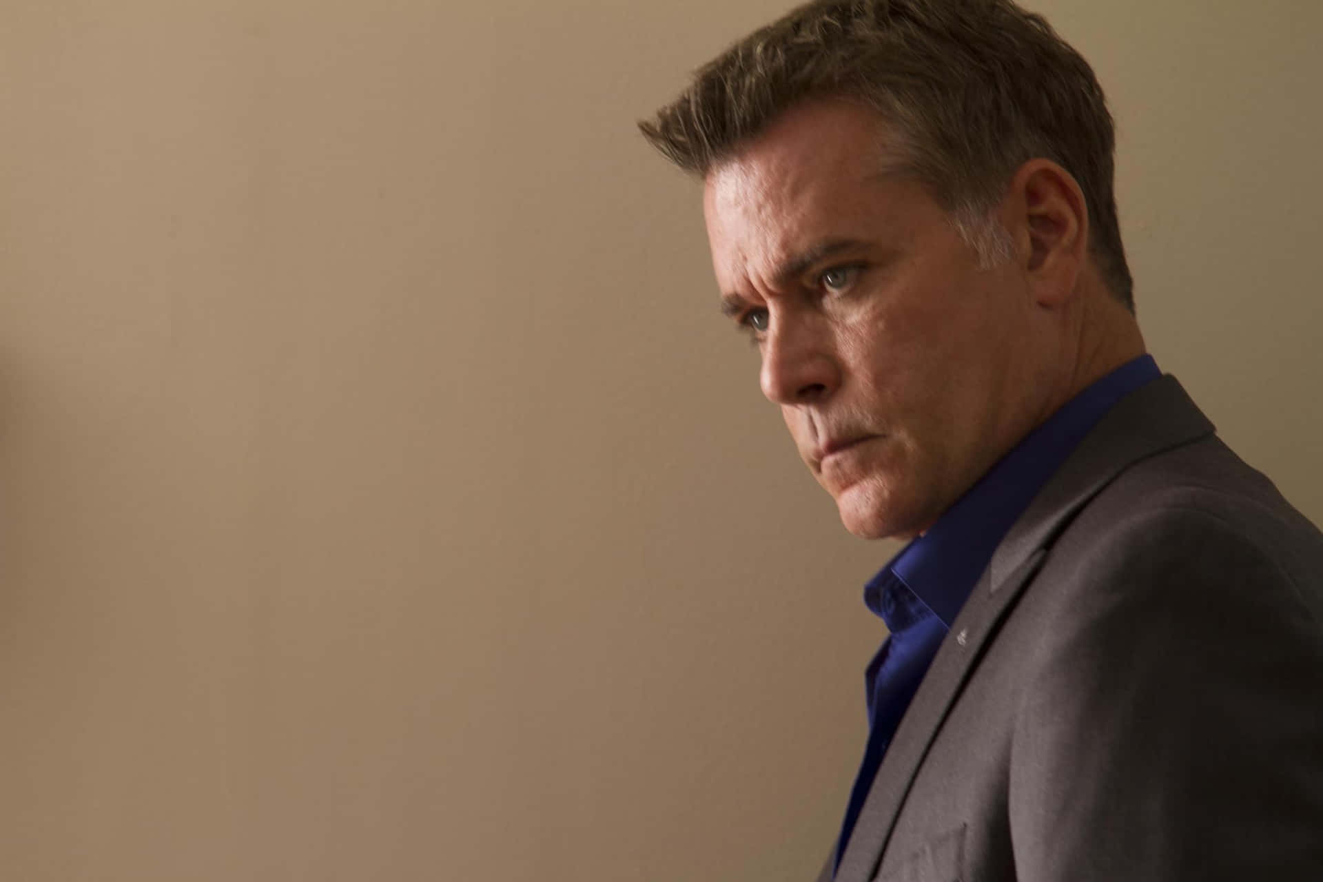 Ray Liotta 桌布 壁紙 — 一名穿著灰色西裝和藍色襯衫的男子，在純米色牆壁前專注地凝視著鏡頭外。