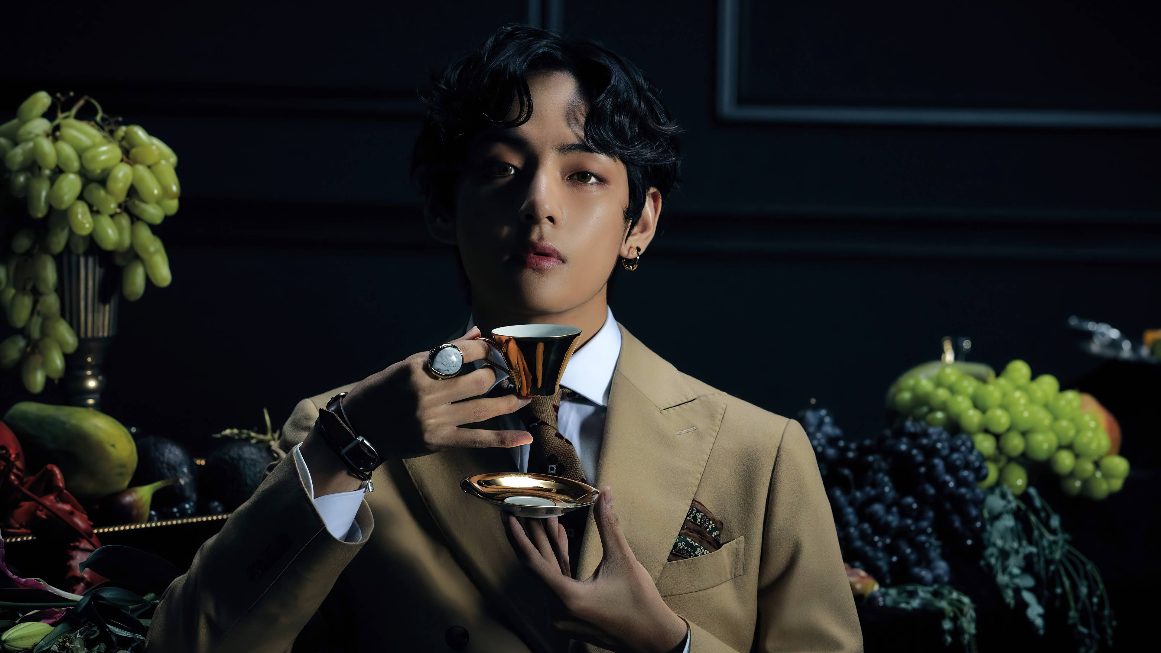 Taehyung《Map Of The Soul: 7》 壁紙 — 一名穿著米色西裝的男子在昏暗的攝影棚中，手持金色杯子，周圍環繞著茂盛的綠色和紫色葡萄。