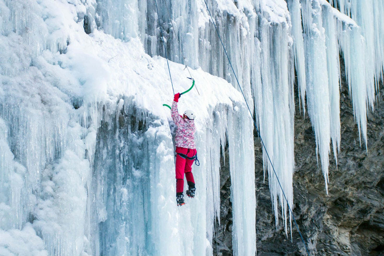 Ice Climbing 桌布 壁紙 — 一名穿著粉紅色裝備的攀岩者在明亮的日光下攀登一座高聳的結冰瀑布，瀑布上掛滿了冰柱。