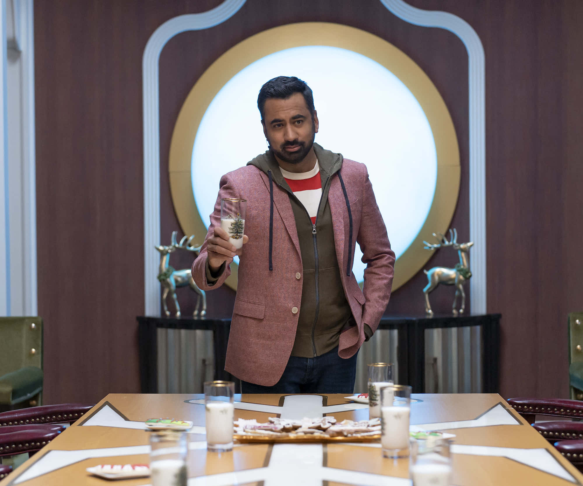 Kal Penn 桌布 壁紙 — 一名穿著粉紅色西裝外套的男子站在現代房間的餐桌後面，手持一杯牛奶，房間內有鹿雕像和發光的圓形牆壁裝飾。