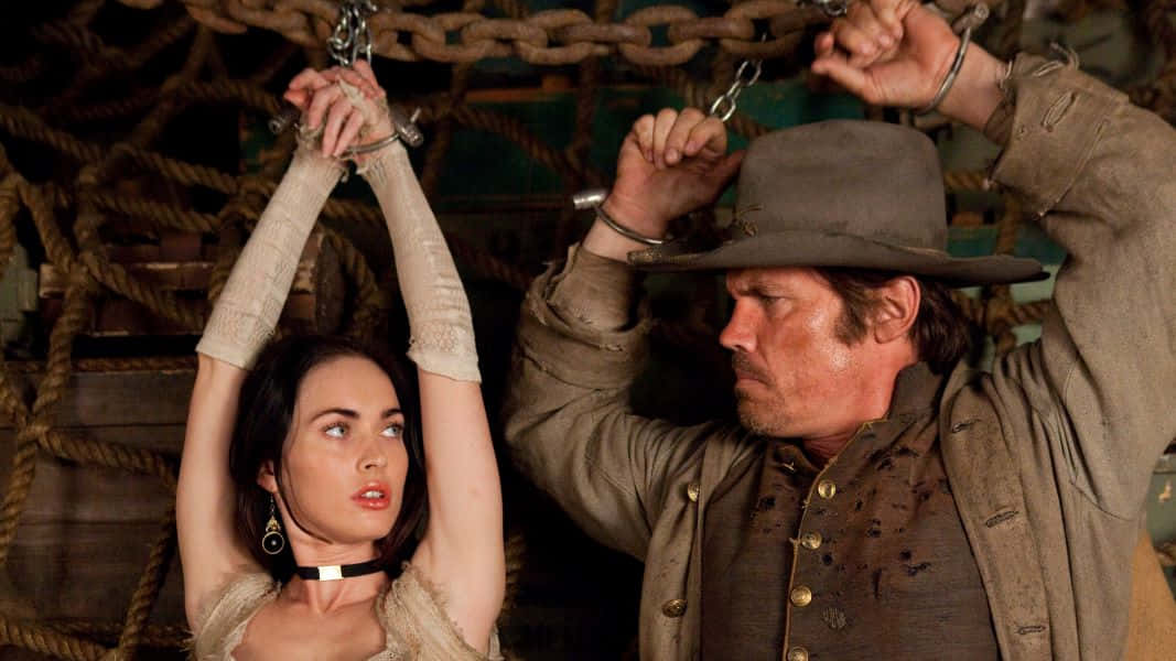 Josh Brolin 桌布 壁紙 — 一名穿著粗獷西部服裝的男子和女子被鎖鏈懸掛在光線昏暗、繩索密佈的船艙中。