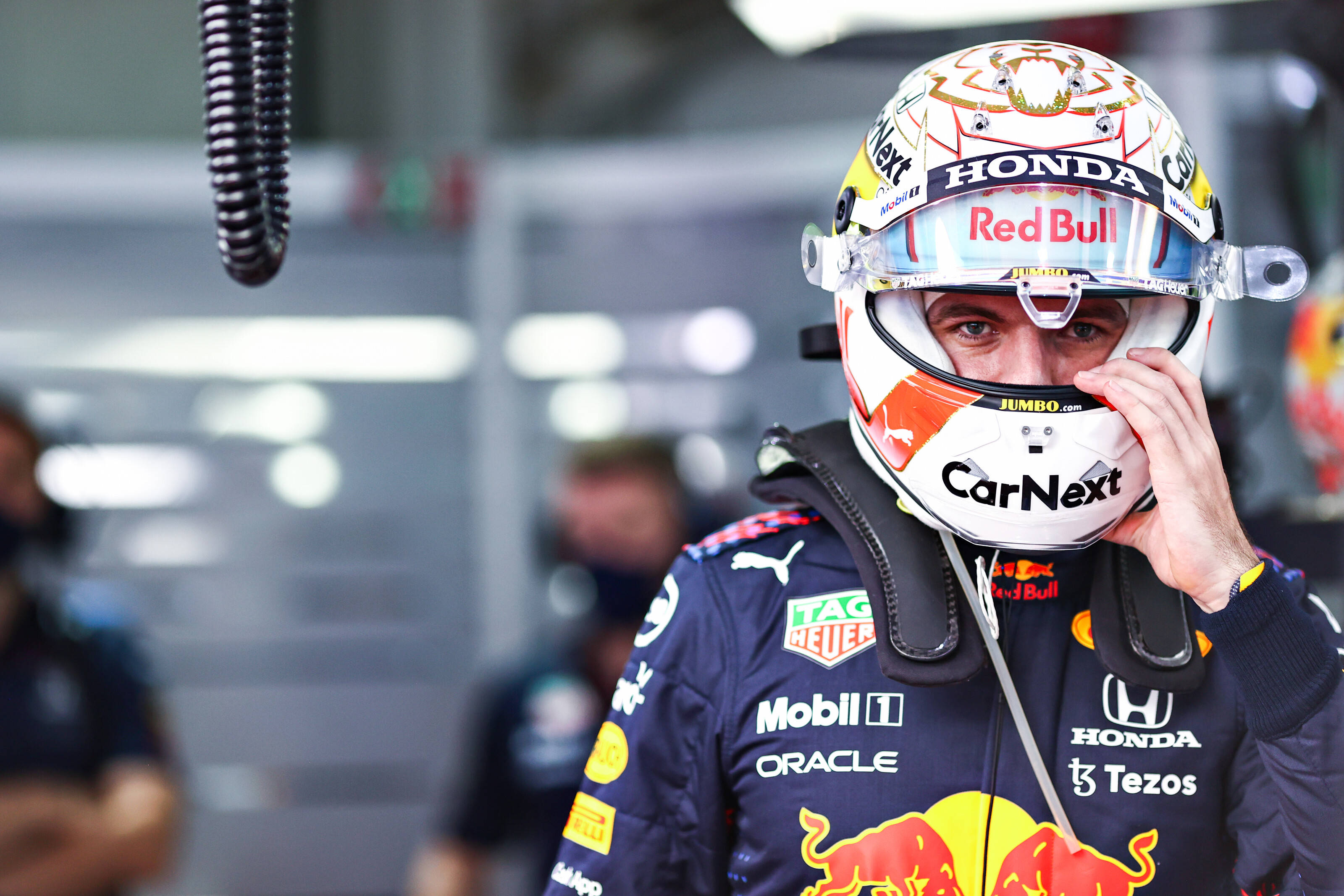 Verstappen 桌布 壁紙 — 一名穿著紅牛賽車服的F1車手在車庫內調整頭盔，背景有模糊的車隊成員。