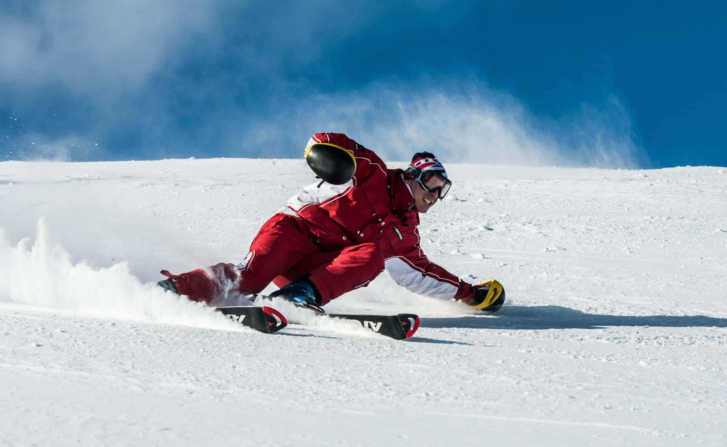 Skiing 桌布 壁紙 — 一名穿著紅色滑雪服的滑雪者在晴朗的藍天下穿過新雪，激起粉雪飛揚。