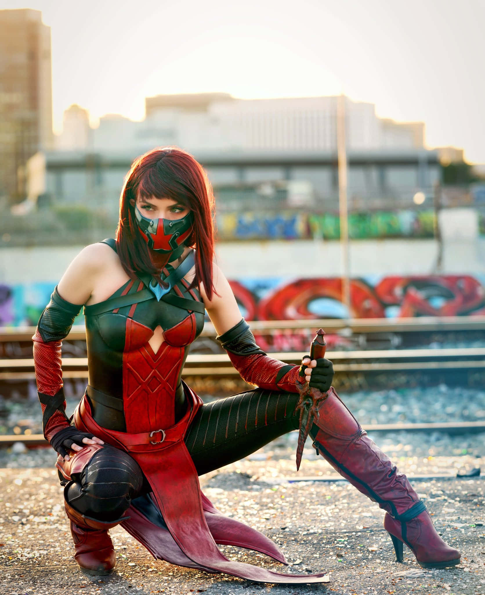 Mortal Kombat Cosplays 桌布 壁紙 — 一名穿著紅黑服裝的角色扮演者在佈滿塗鴉的火車軌道附近的礫石上蹲下，背景是城市天際線。